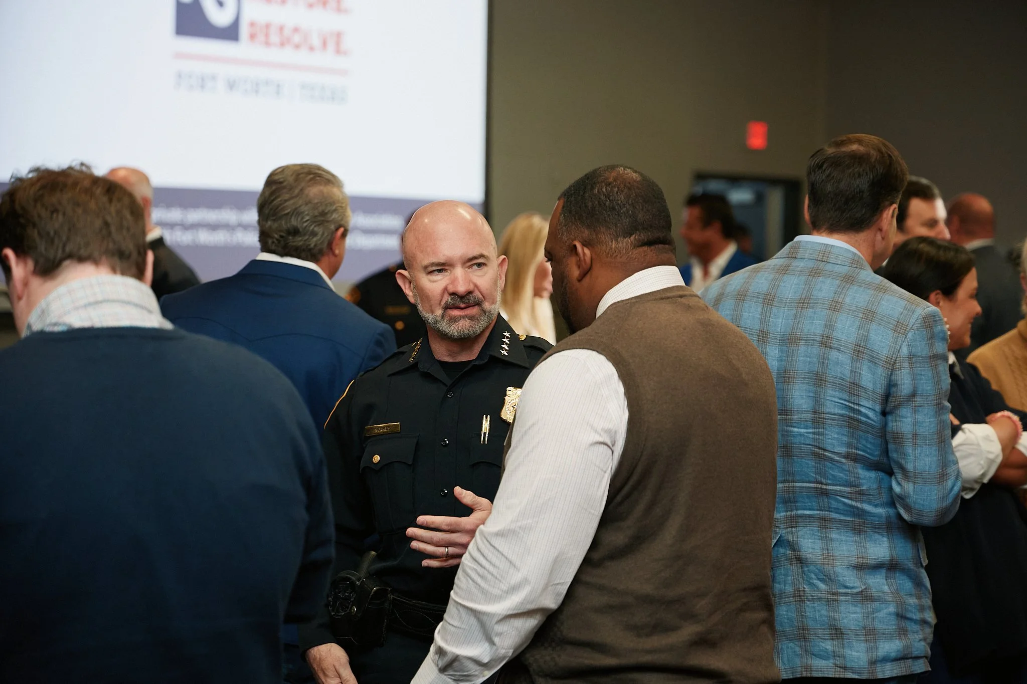 A police officer talking to a man at a conference or event with a large screen in the background that has part of a logo and text, with several other people in conversation.
