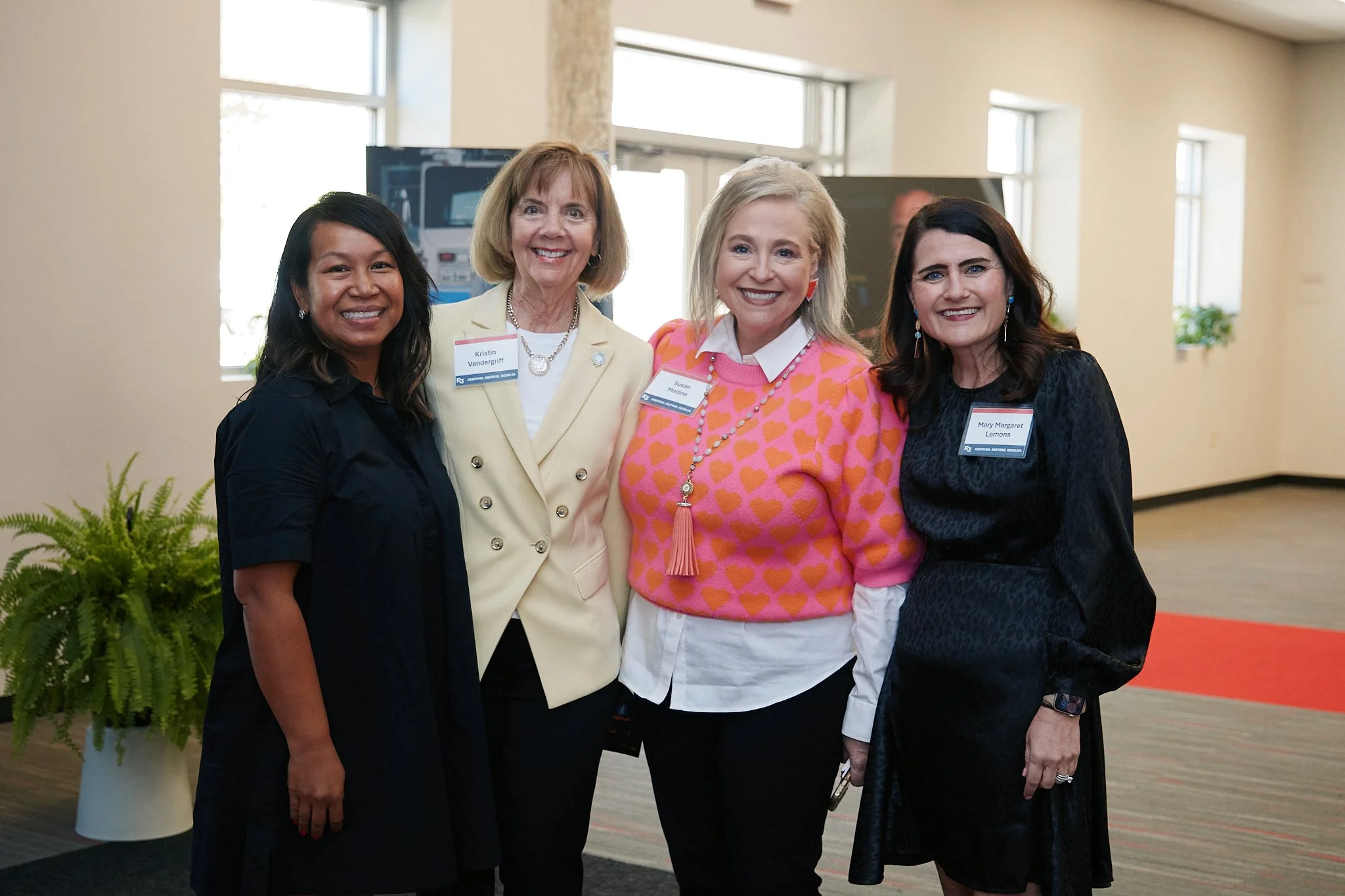 Four smiling women standing together indoors at a professional event, wearing name tags and colorful clothing.