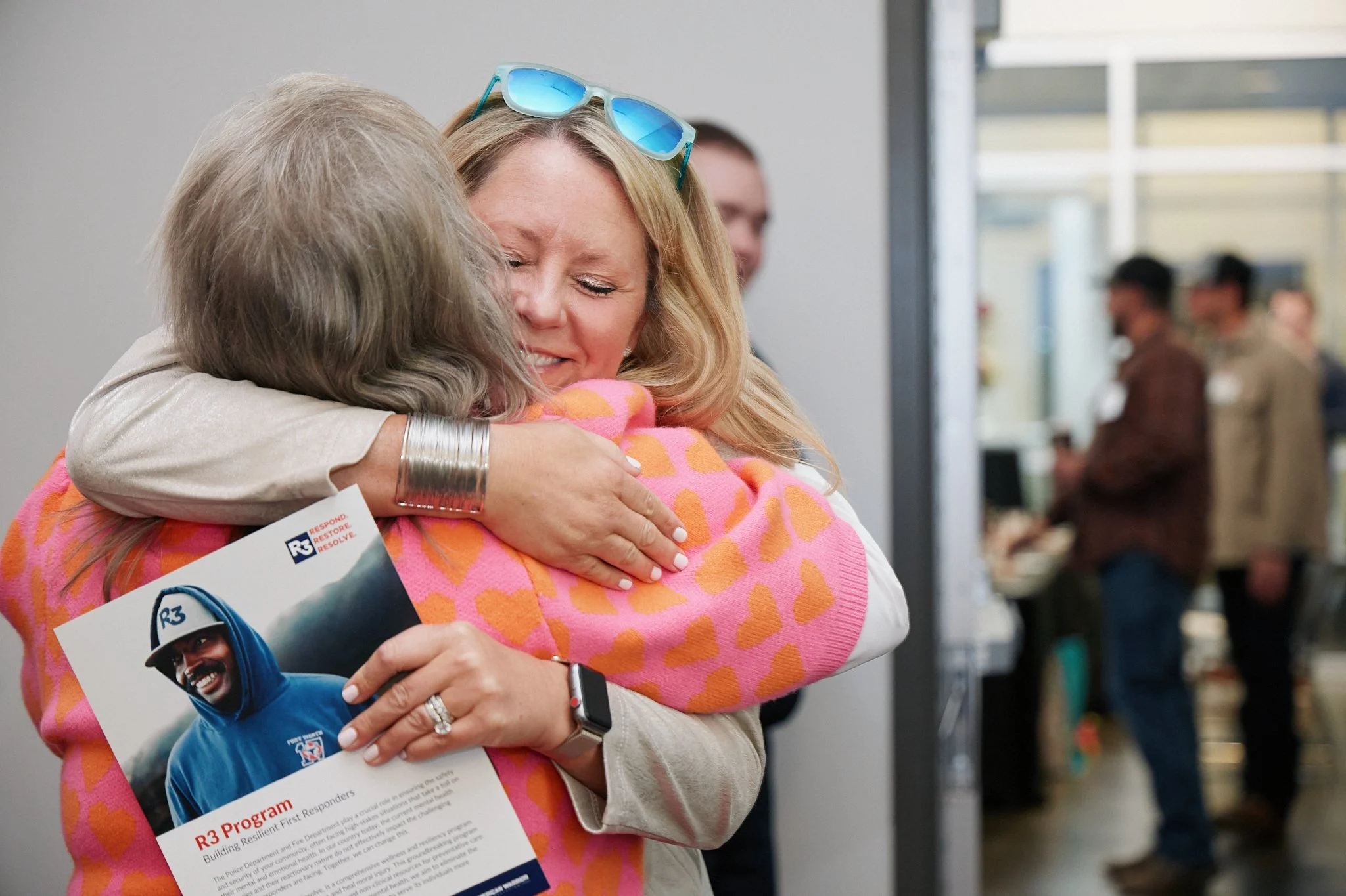 Two women embrace each other warmly at an indoor event. One woman, with blonde hair, is wearing sunglasses on her head and a pink sweater with orange hearts. The other woman, with gray hair, is holding a brochure or flyer. In the background, there ar