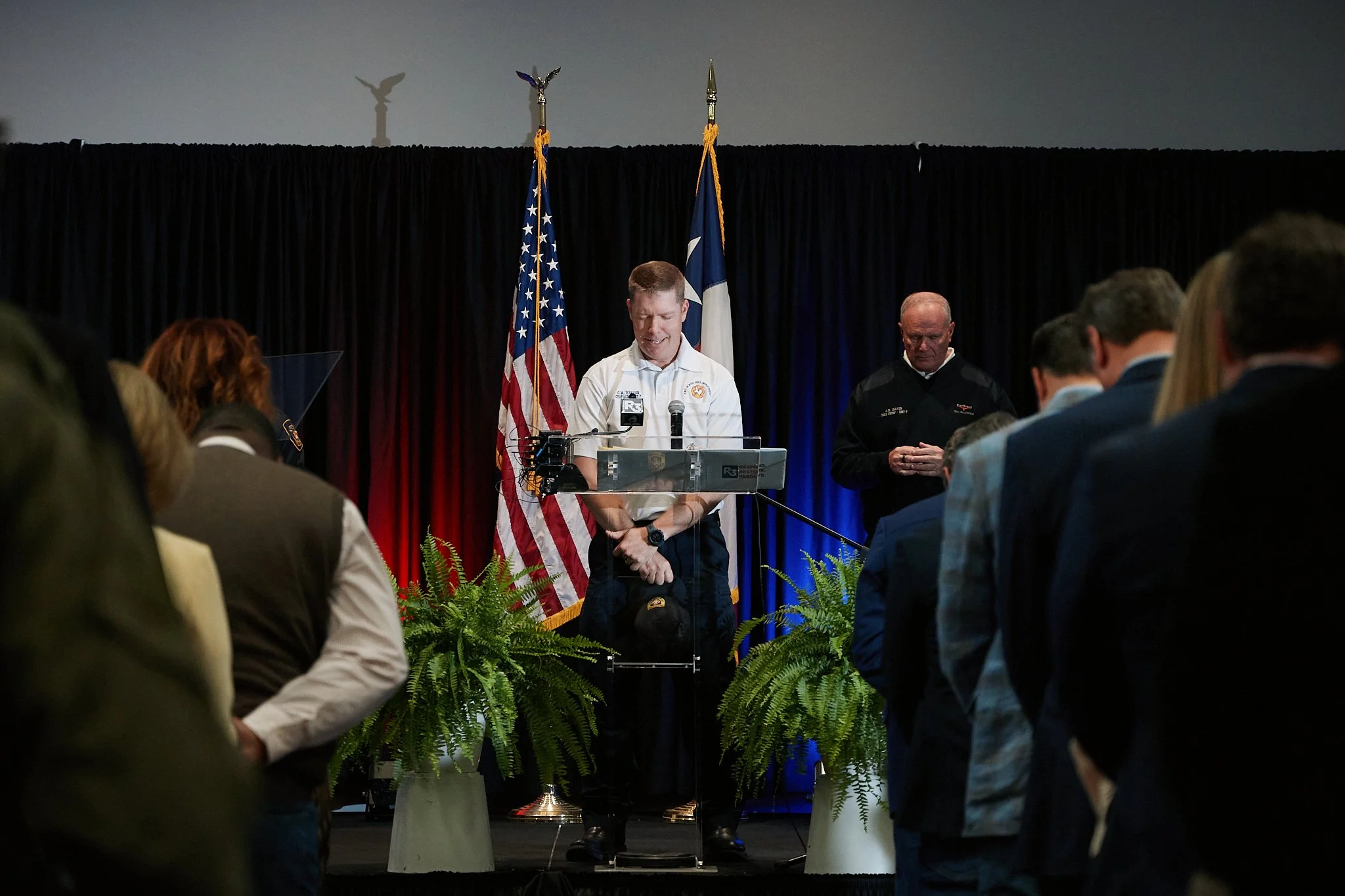 A man in a white shirt standing at a podium with two microphones in front of American and state flags, with people gathered in the audience and a dark curtain backdrop.