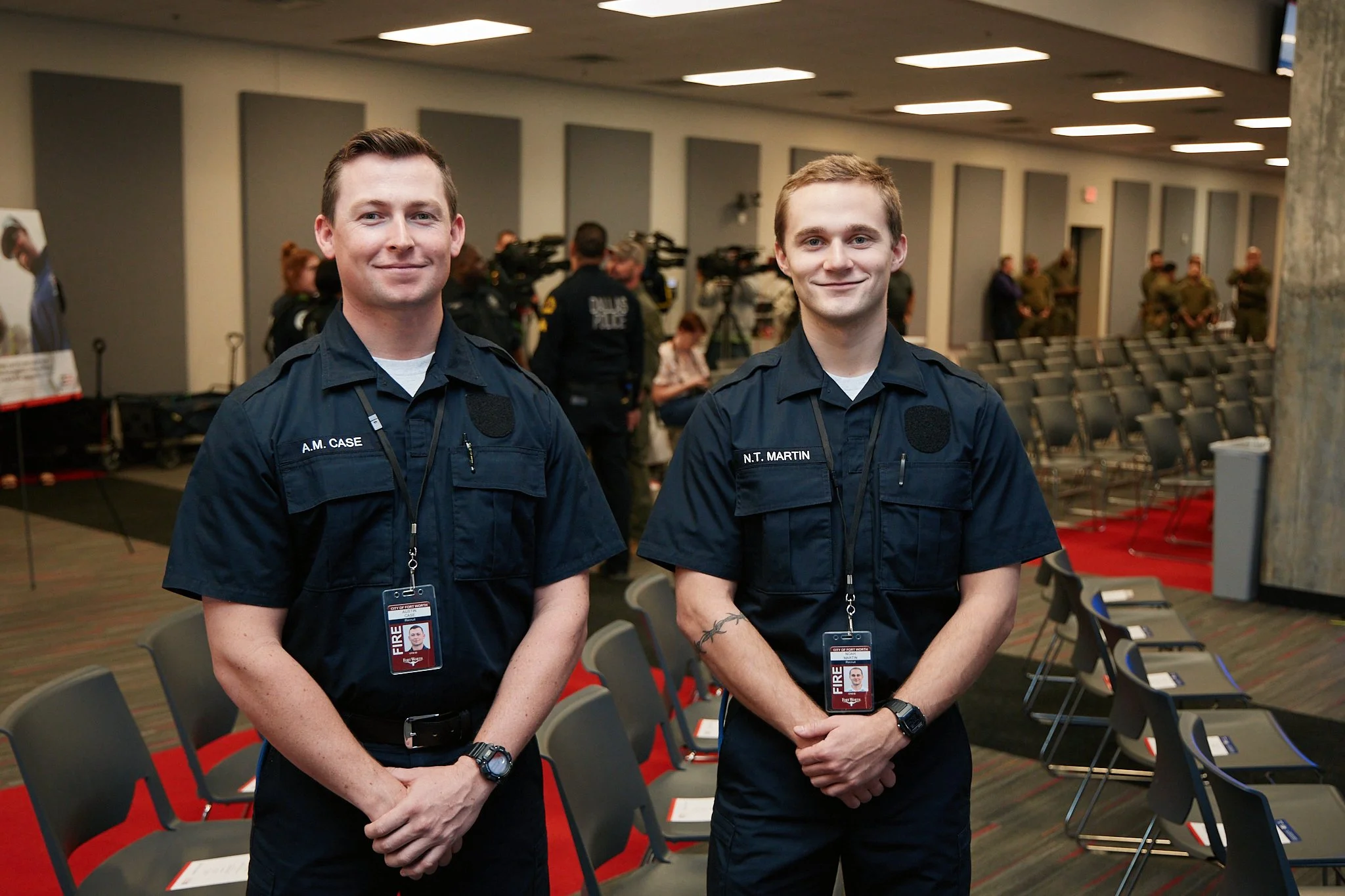 Two young men in navy uniforms with name tags and fire badges standing in a conference room with chairs, microphone cables, and a background of cameras and other people.