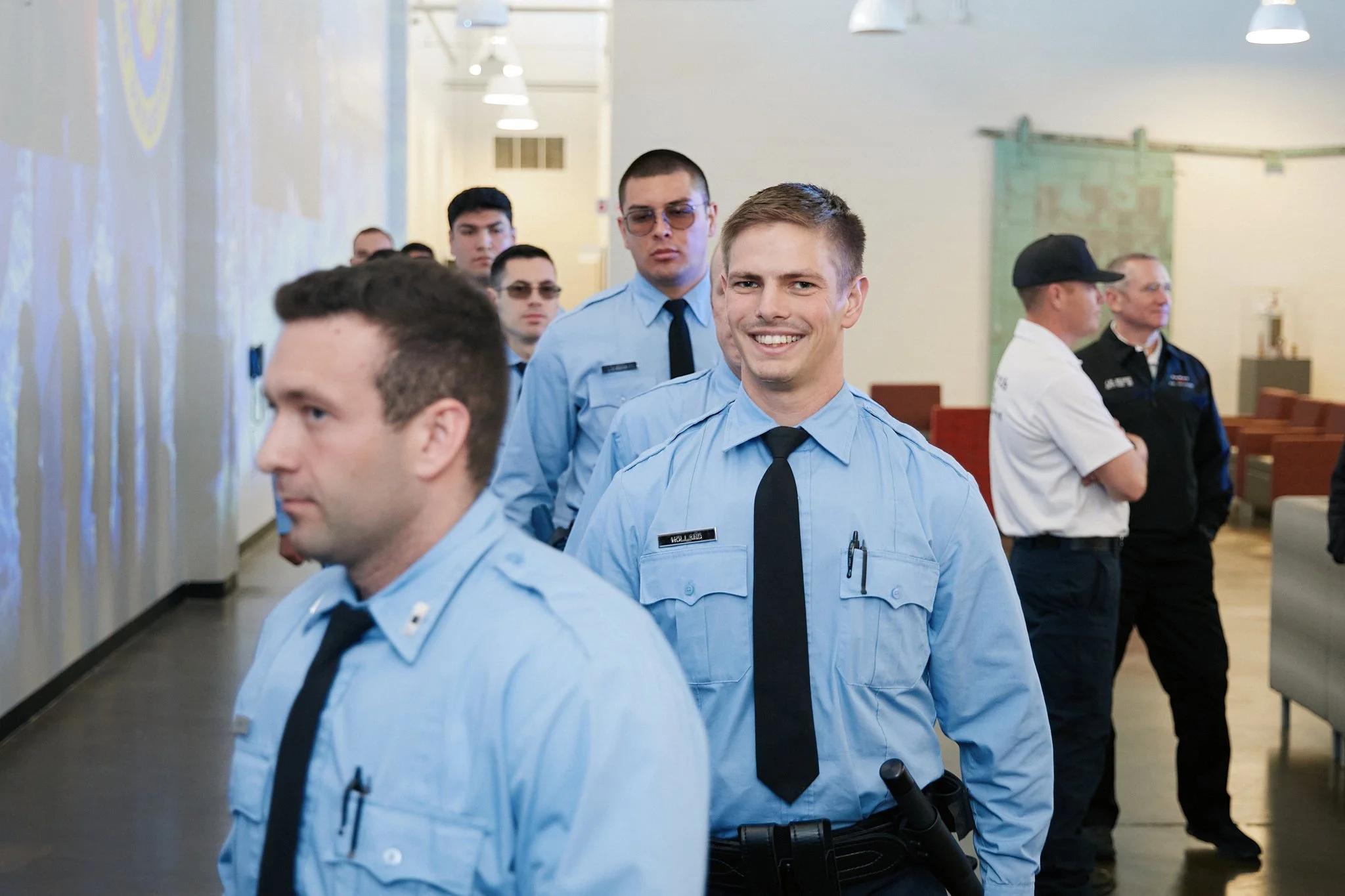 Group of uniformed military personnel in a line, with a smiling man at the front, walking through an indoor area.