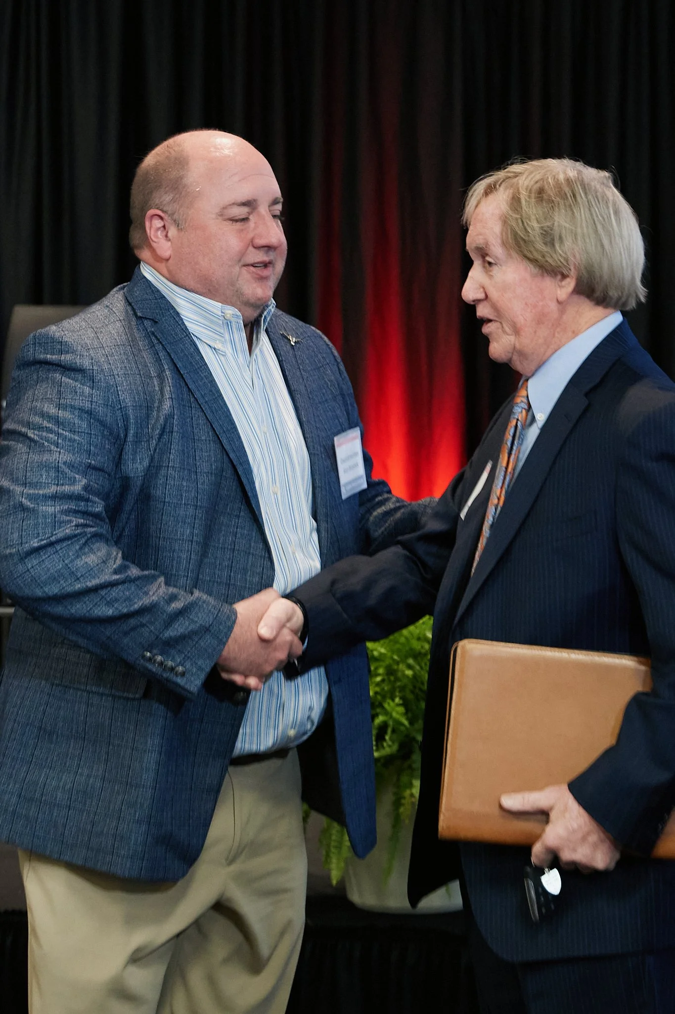 Two men in business attire shaking hands at a formal event, with a man on the right holding a brown folder, in front of a black curtain with red lighting.