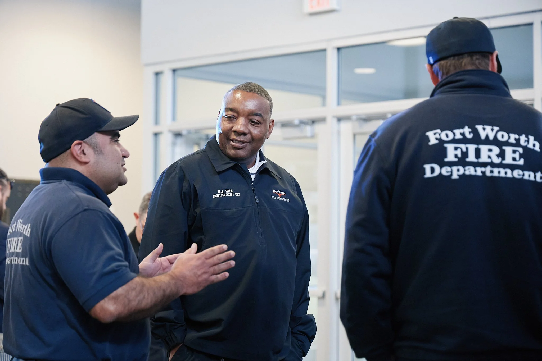 Three men in matching navy blue Fort Worth Fire Department jackets and hats engaged in conversation indoors, with a glass door and people in the background.