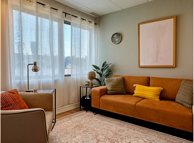 Living room with a light green wall, a mustard yellow sofa with green and yellow pillows, a beige armchair with an orange pillow, a black side table with a plant and decorative items, a modern black clock on the wall, a framed line drawing, sheer white curtains, and a beige carpet.