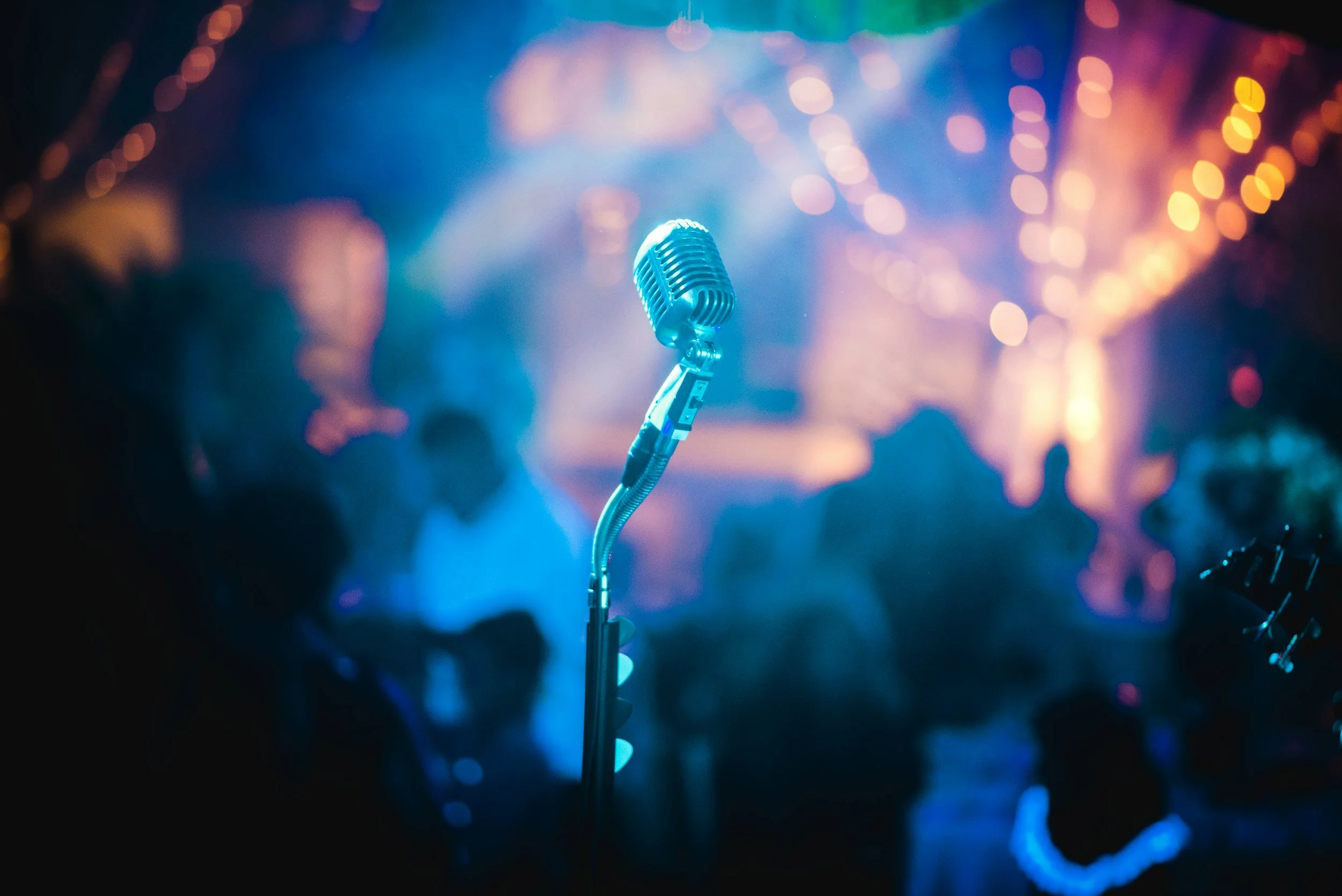 A vintage microphone on a stand in front of a blurred crowd at a dimly lit stage with colorful lights.