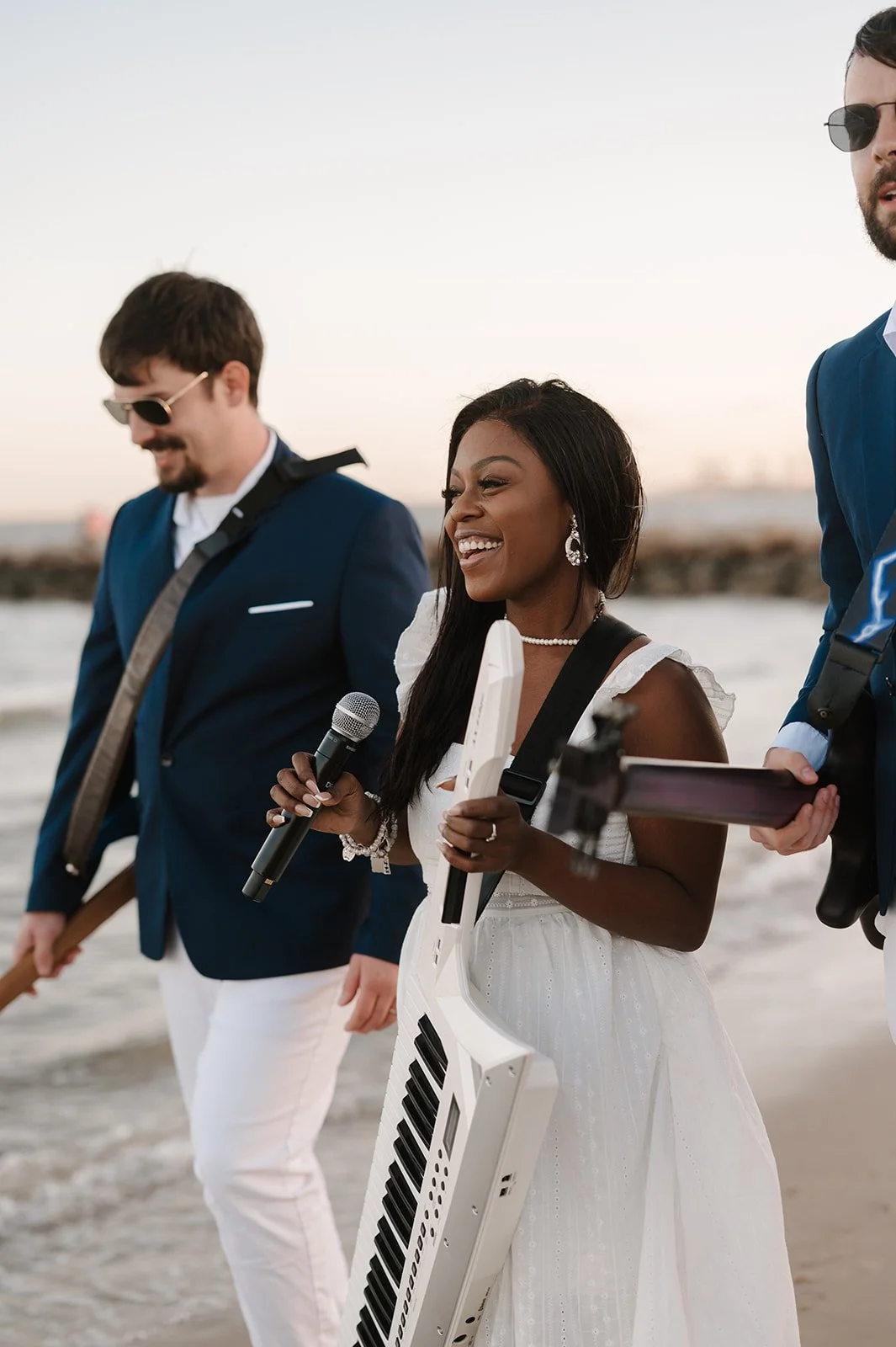A group of three musicians walking on the beach, including a woman with a keytar, holding a microphone, singing, and smiling, with a man in a suit and two other men with musical equipment.