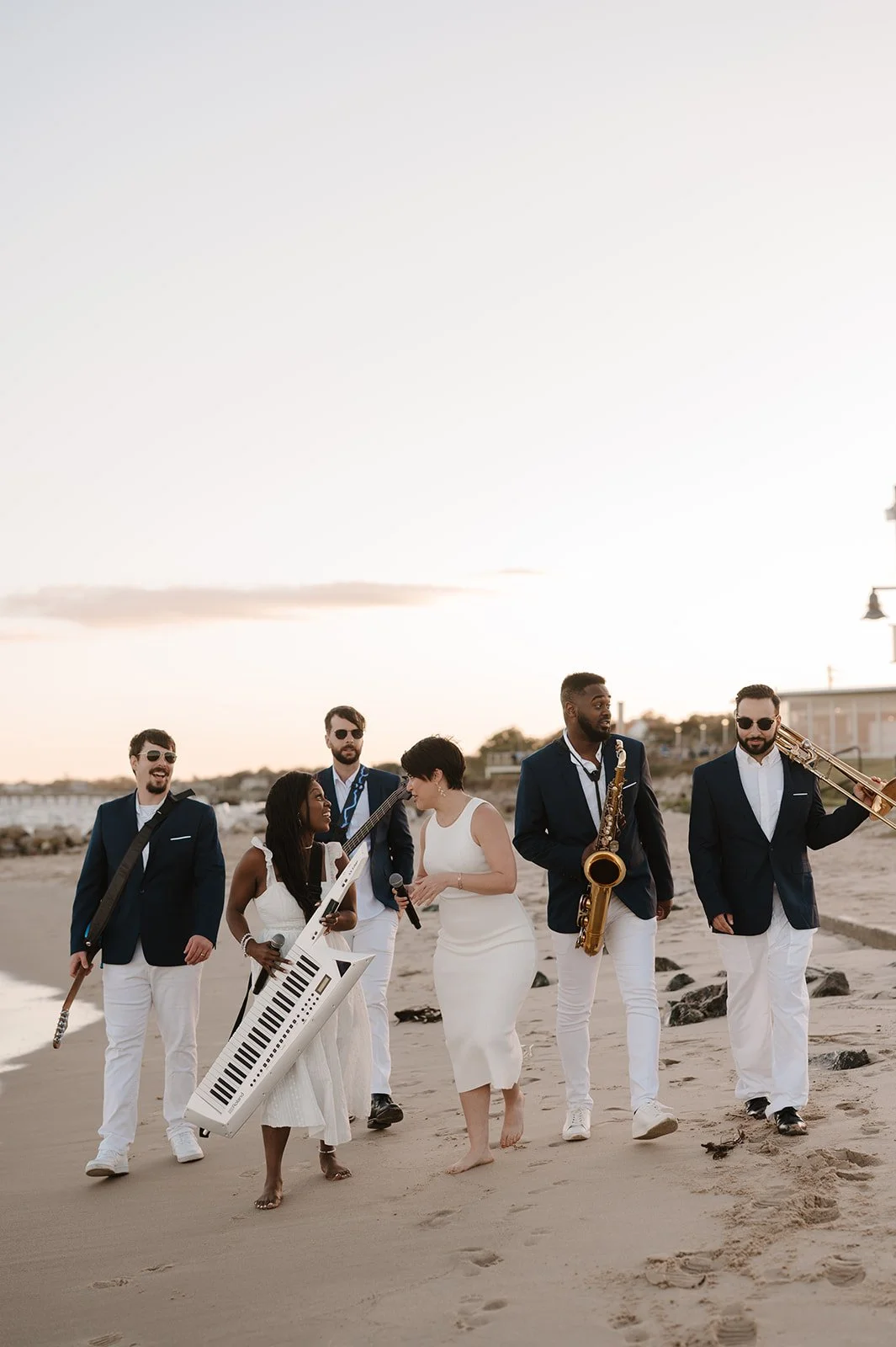 A band of six musicians walking on a beach at sunset, dressed in formal attire, holding musical instruments including a keytar, electric guitar, saxophone, and trombone.