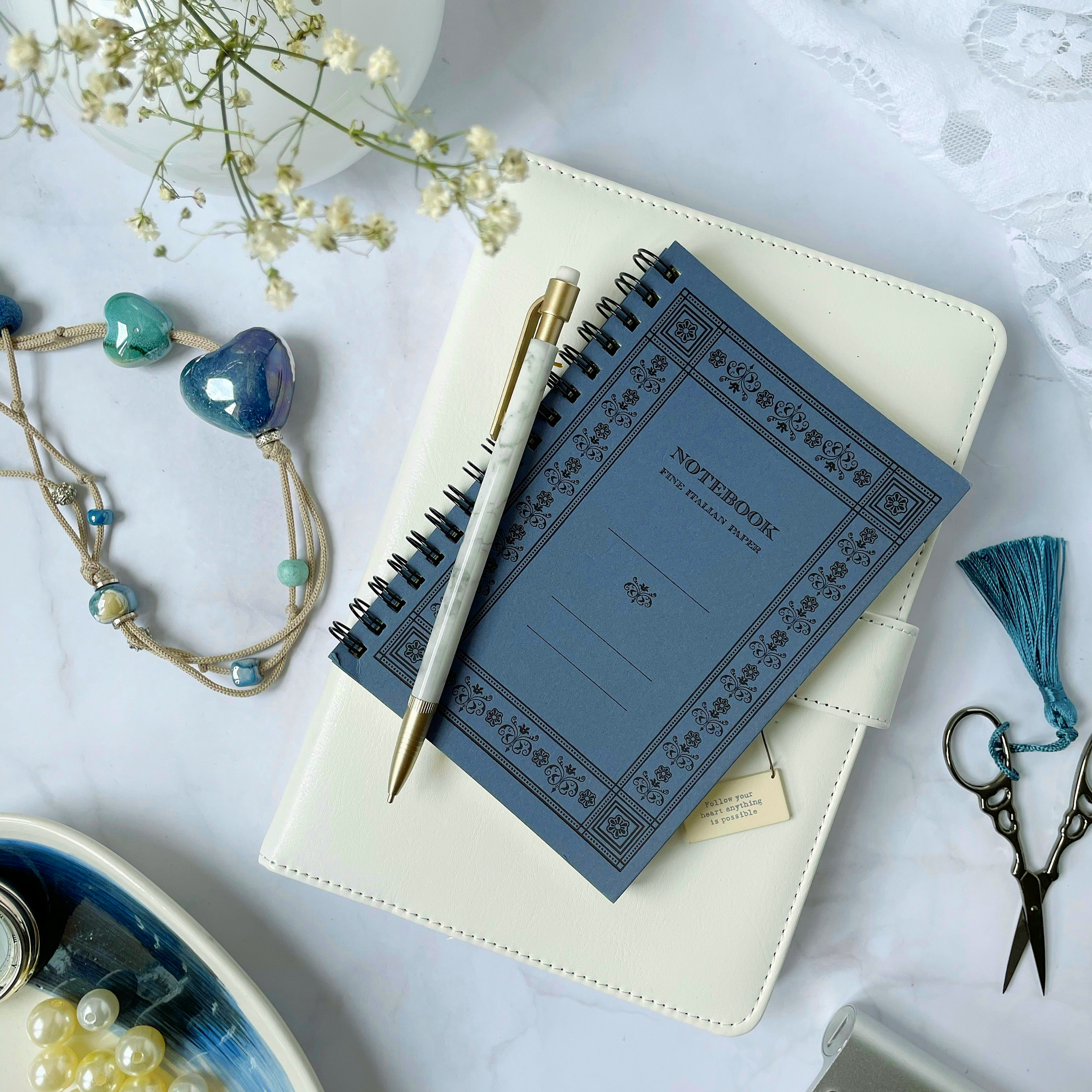 Blue spiral notebook and pen on a cream planner, with a beaded necklace, blue heart pendant and teal tassel with scissors.