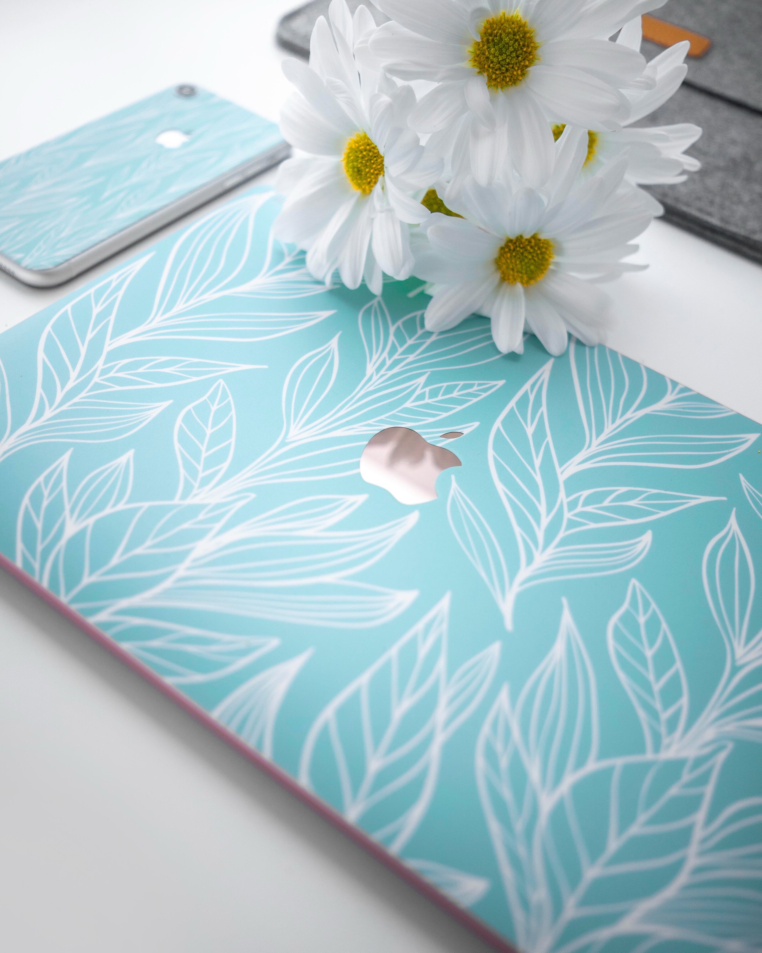 Close-up of a turquoise laptop with a white leaf pattern, matching phone case and white daisies resting on top