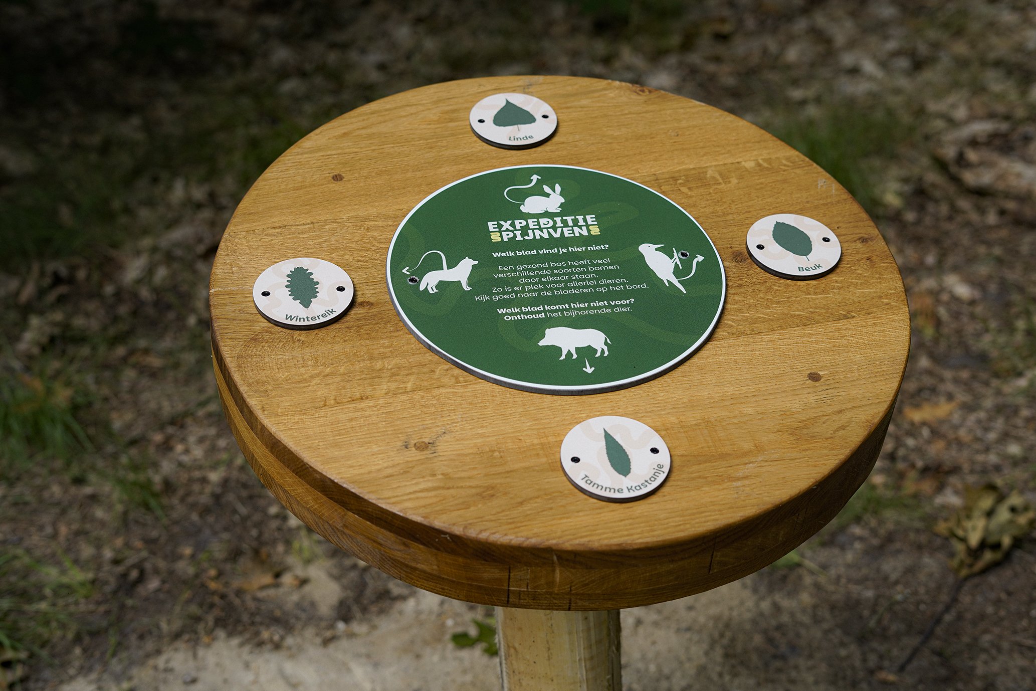 Round wooden information table with a green circular sign in the center and four smaller oval signs around it, set outdoors on a wooden post. The sign has text in Dutch and images of animals for a nature trail.