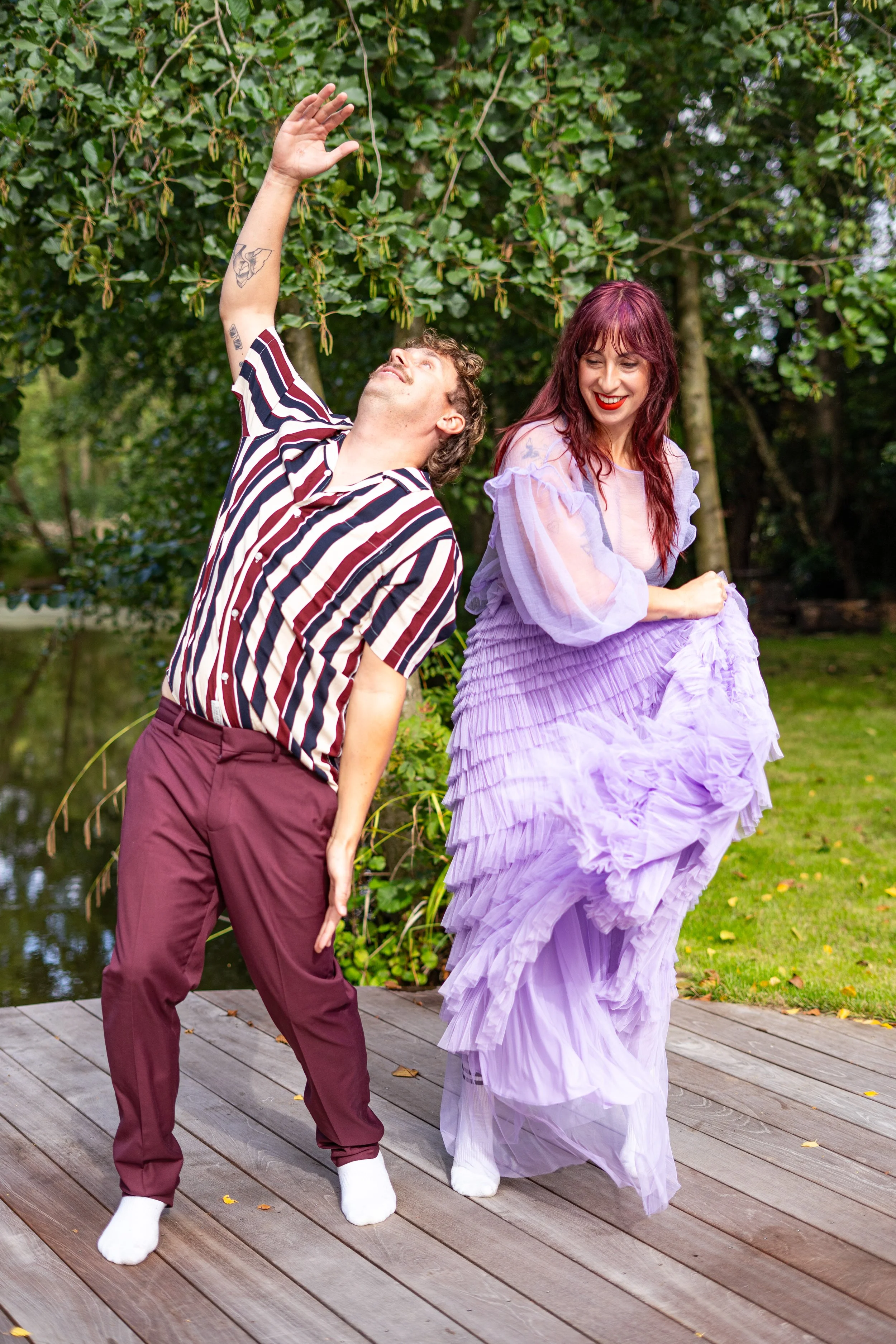 Two people, a man and a woman, standing on a wooden deck near a pond with trees in the background, dancing playfully. The man is wearing a striped shirt and maroon pants, while the woman is wearing a light purple ruffled dress with sheer sleeves. Both are wearing white socks and are smiling, with the man raising one arm and the woman looking at him.
