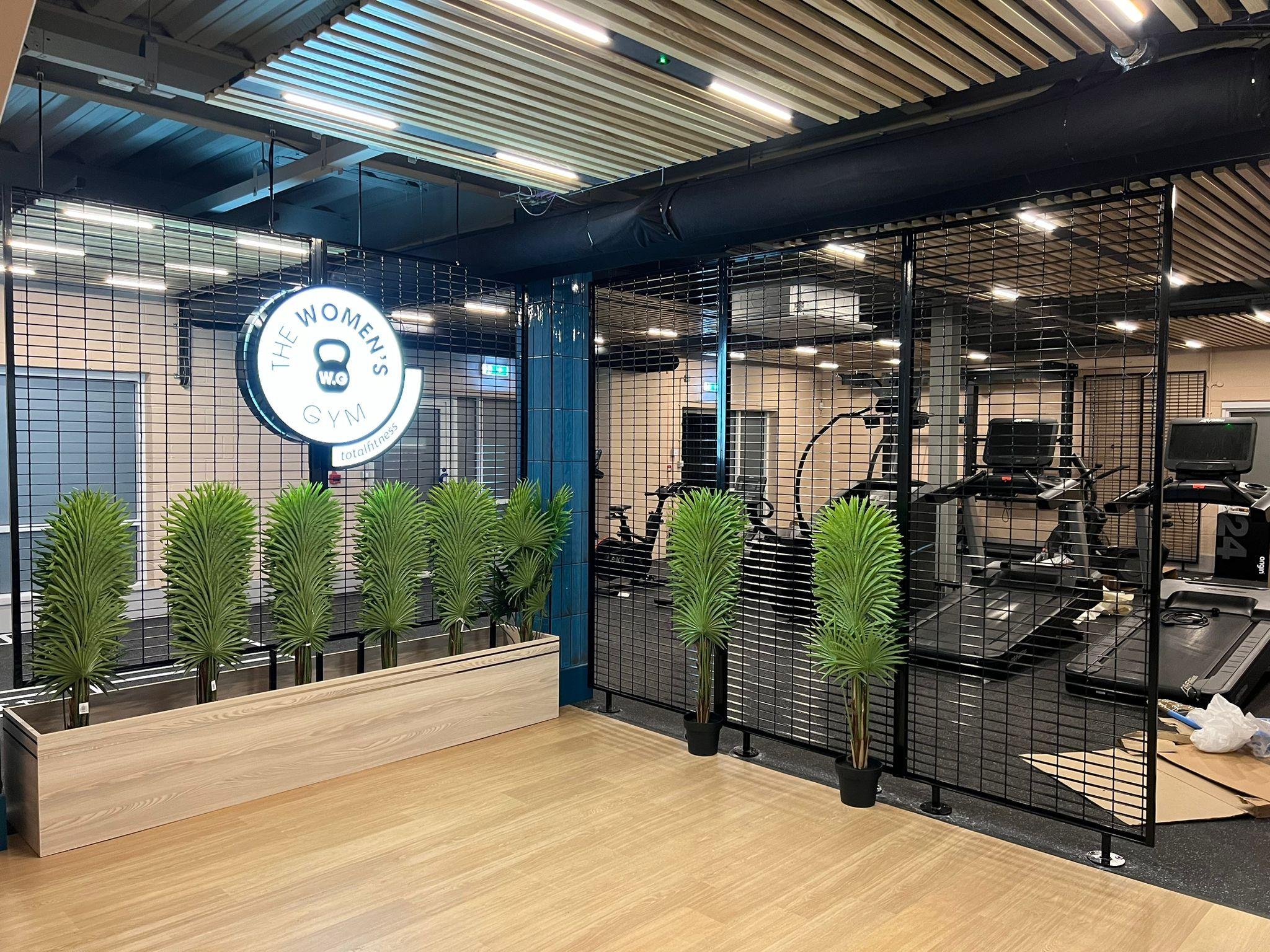 Entrance to a women's gym with a sign that reads 'The Women's Gym' and a logo featuring a kettlebell, with a row of tall green plants in a wooden planter in front and gym equipment visible inside.