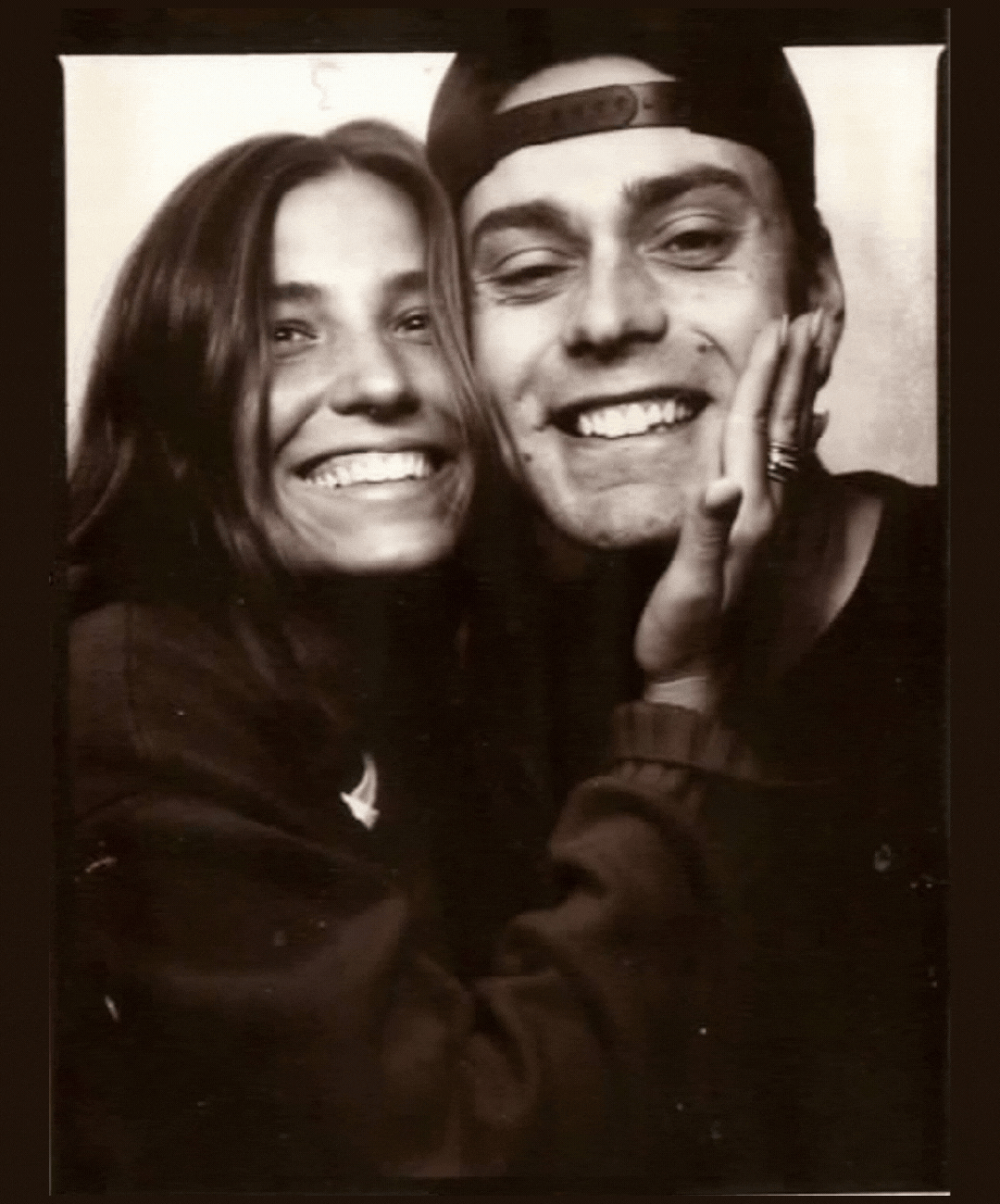 Black and white photo of a smiling woman with long hair and a man with a backward cap, with the woman's hand on the man's cheek.
