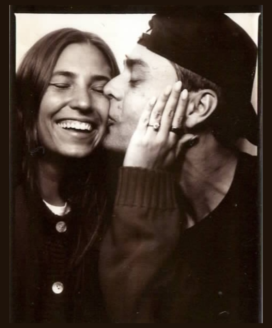 A black and white photo of a man and woman sharing a kiss, with the woman smiling and holding the man's face, showing her wedding ring and engagement ring.