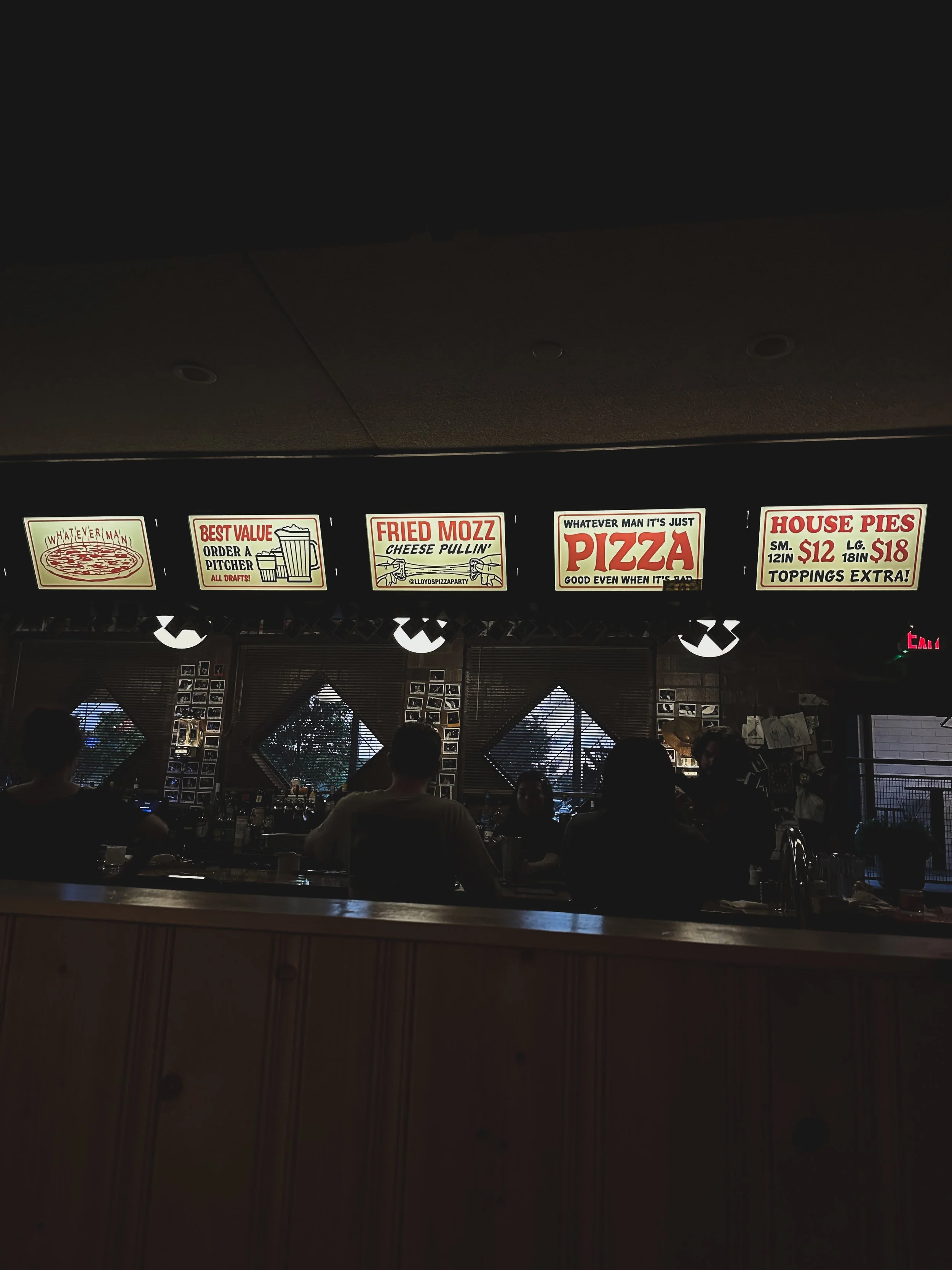 Bar counter with illuminated menu signs displaying pizza and house pies prices, and people sitting at the bar with windows in background.
