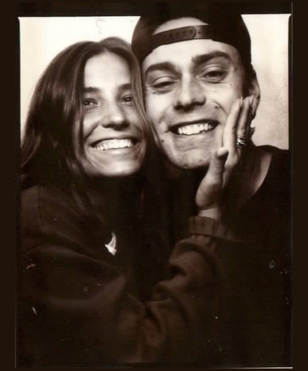 Black and white photo of two people smiling, a woman with long hair and a man wearing a backwards cap, with the woman's hand on the man's face.
