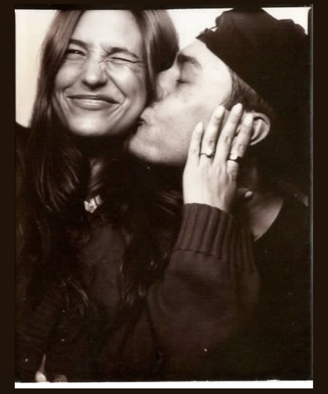 A woman with long hair and a big smile is about to kiss a man wearing a beanie hat; she is holding his face with her hands.