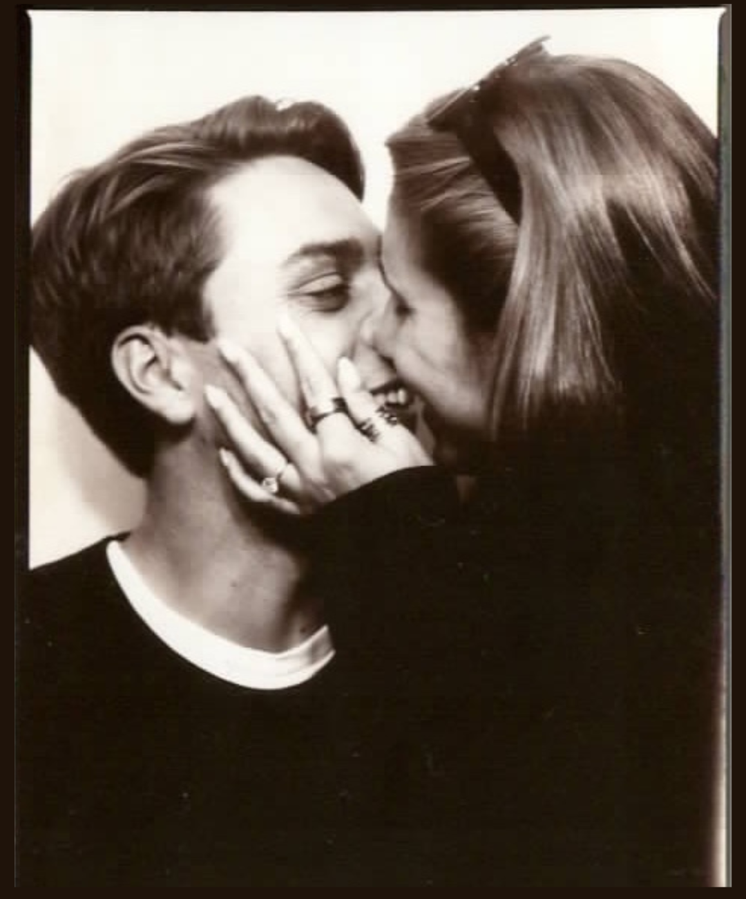 Black and white photo of a young man and woman smiling as they share a kiss, with the woman's hand gently holding the man's face and her fingers adorned with rings.