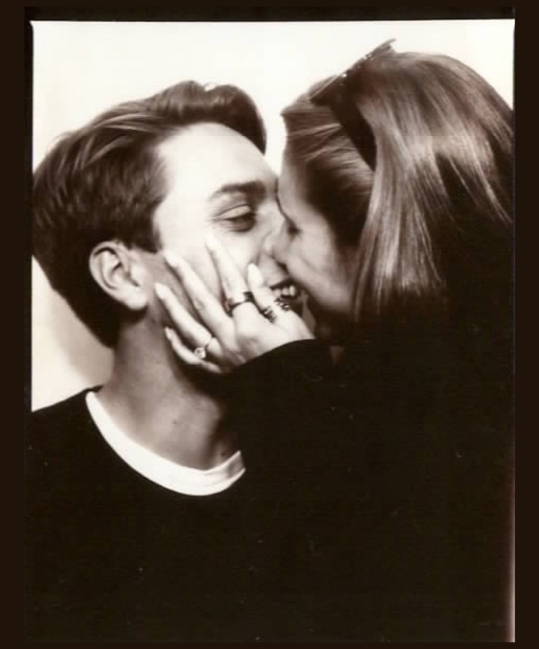 A black and white photograph of a couple sharing a kiss, with the woman holding the man's face, both wearing dark clothing.