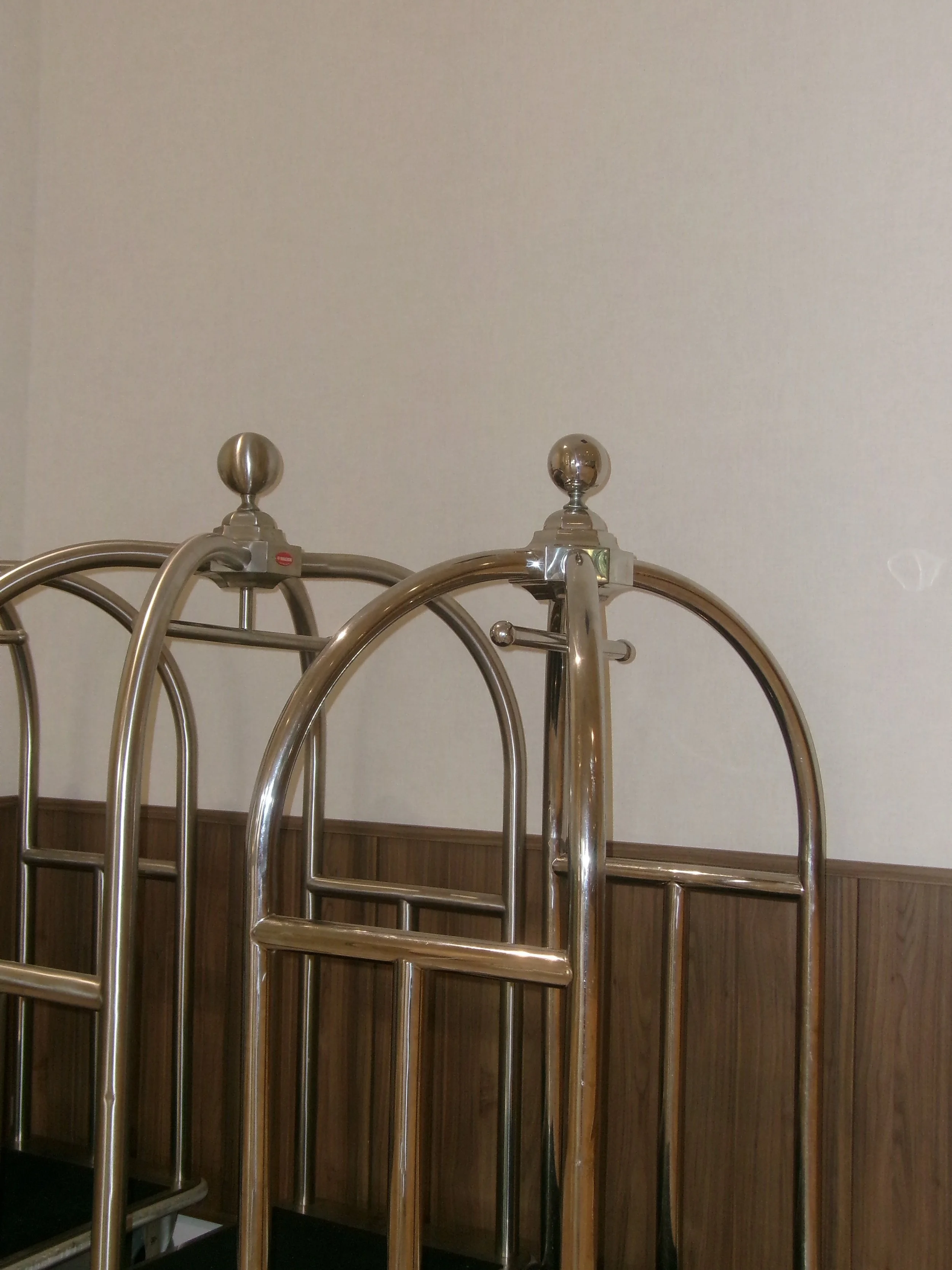 A brass luggage cart with a metal ball top on the handle, positioned in front of a light-colored wall and wooden paneling.