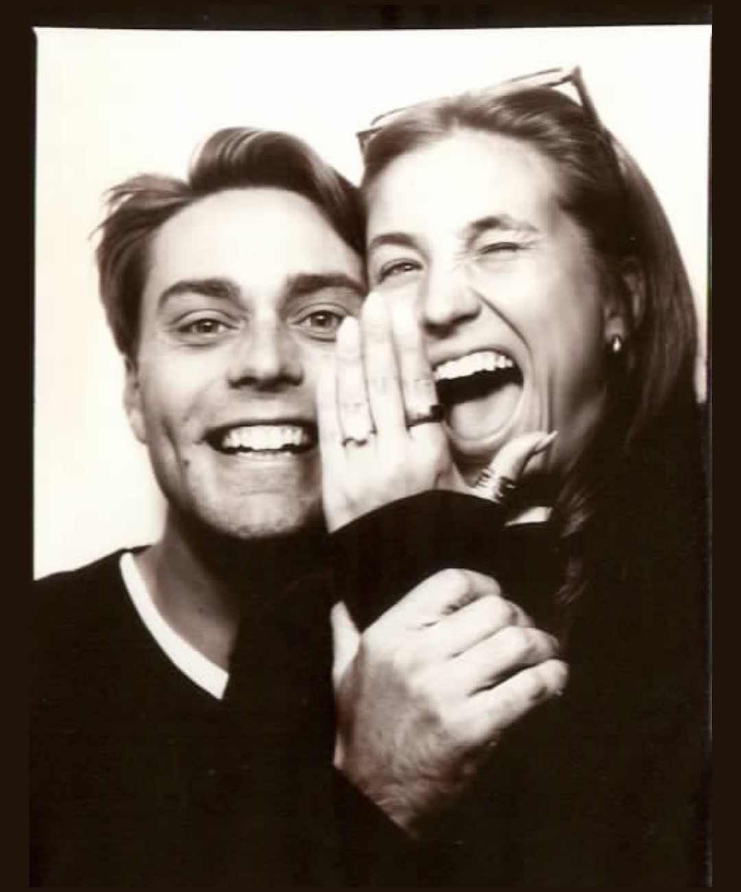 Black and white photo of a happy couple, the man has a broad smile and the woman is playfully whispering or shouting with her hand near his ear, both smiling and showing excitement.