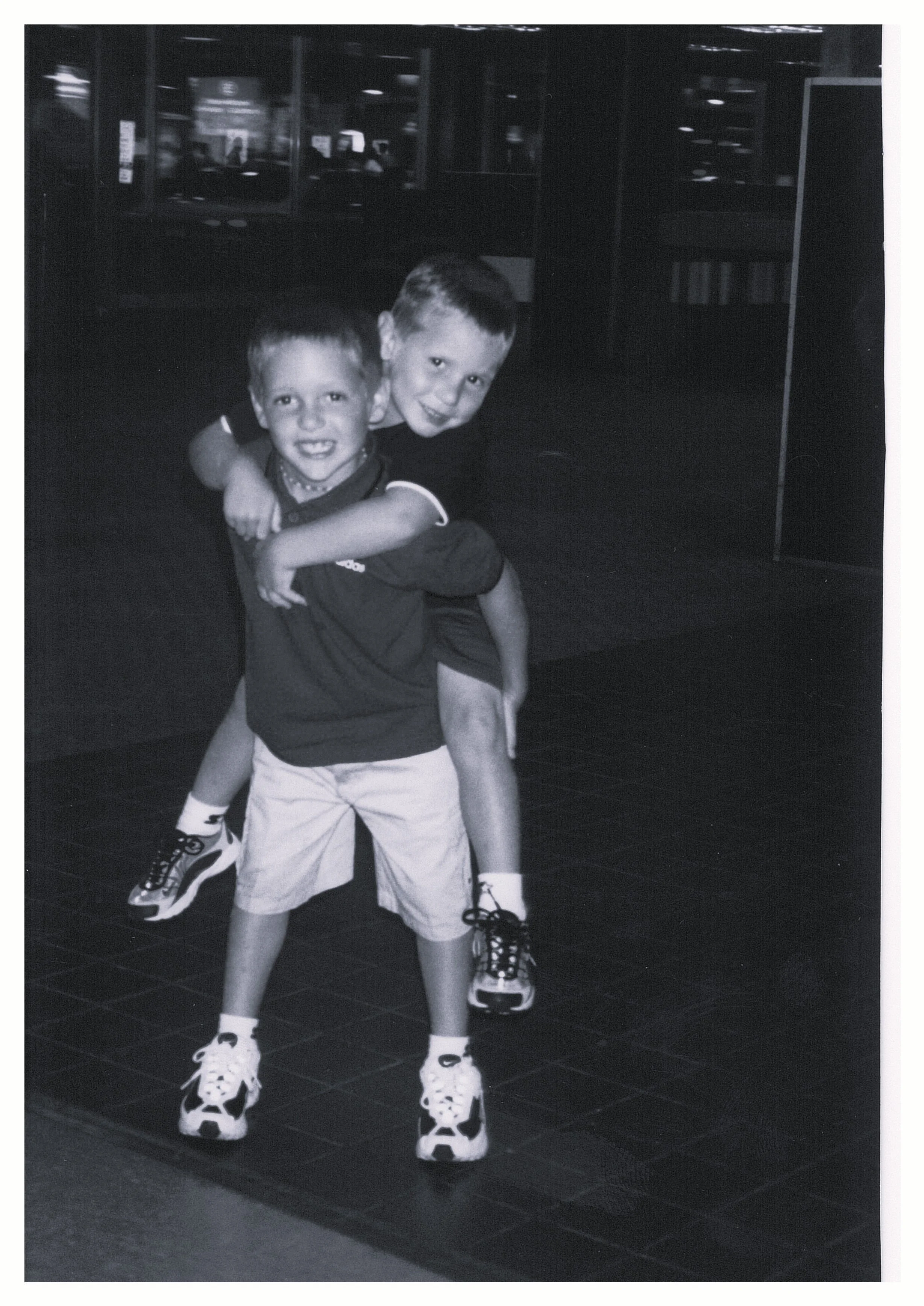 Young man with dark curly hair making a peace sign while sweeping a kitchen floor with a broom and dustpan, some spilled debris on the floor.