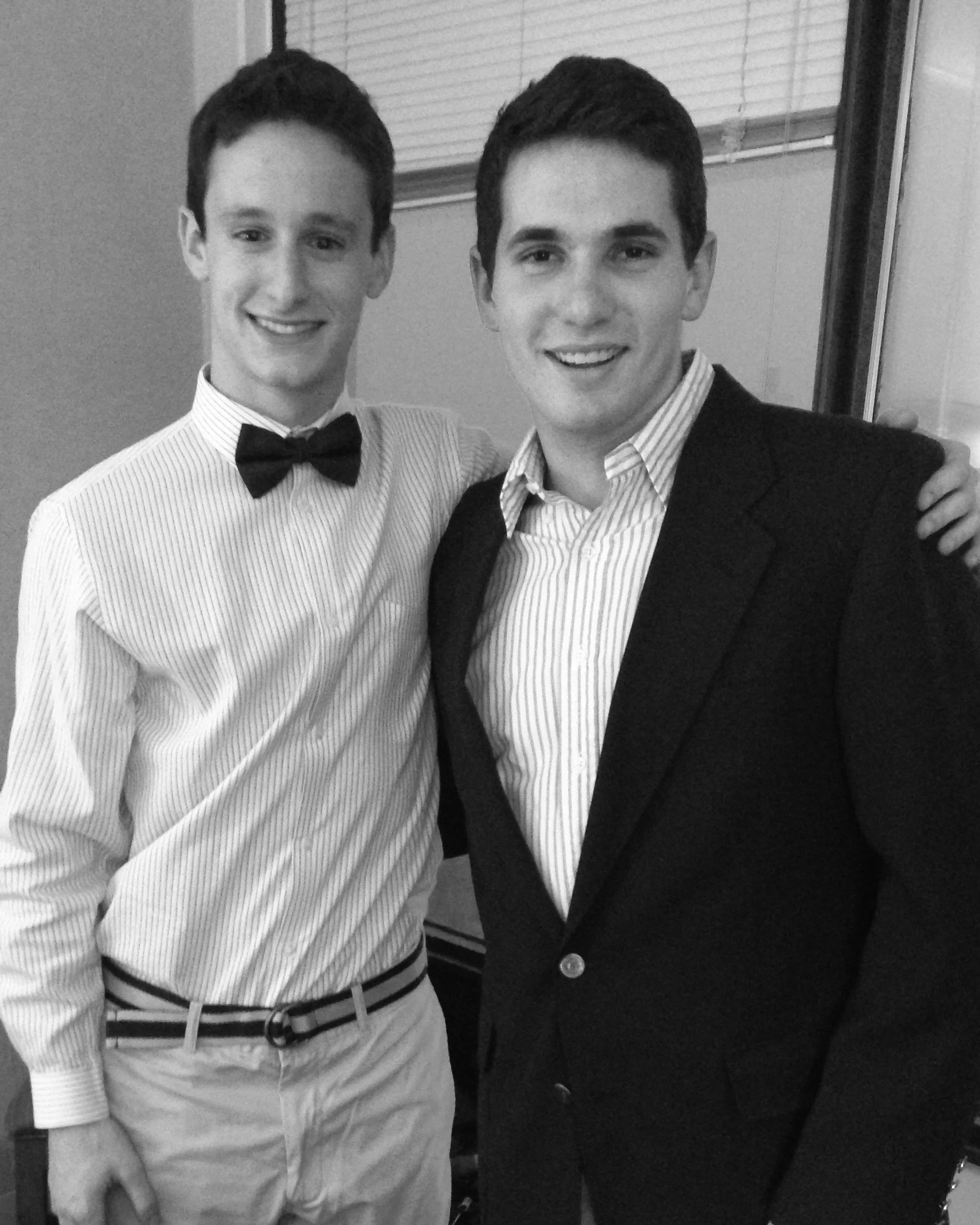 A young woman and man standing in a parking lot at night. The woman wears sunglasses, a black jacket, a white crop top, and a long denim skirt. The man holds a stereo speaker and a boombox. They are surrounded by parked cars, and a lit sign reads 'TICKETS' in the background.