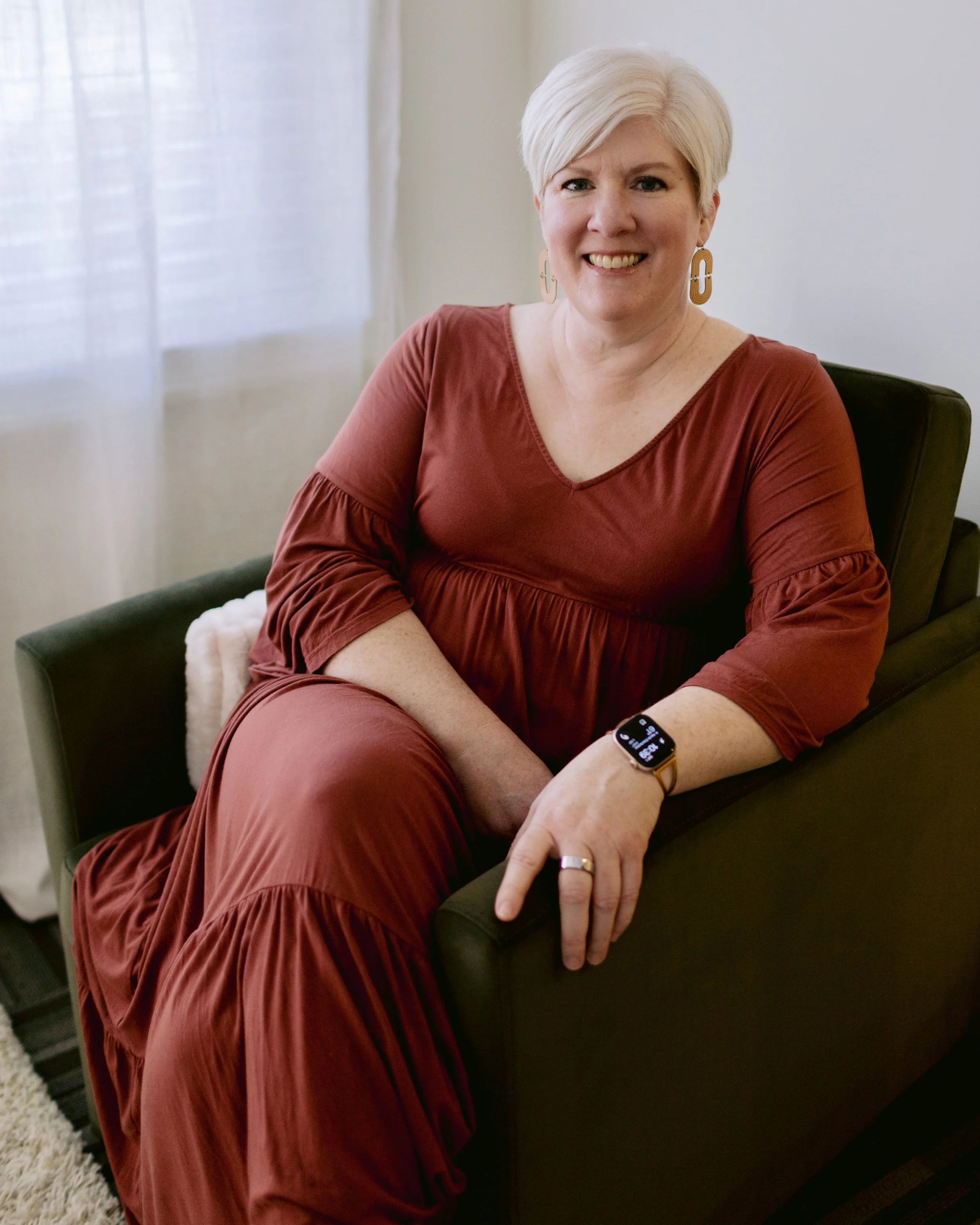 A woman with short, platinum blonde hair, wearing a rust-colored dress, smiling and pointing to her smartwatch on her left wrist, sitting on a dark-colored chair in a room with white curtains.