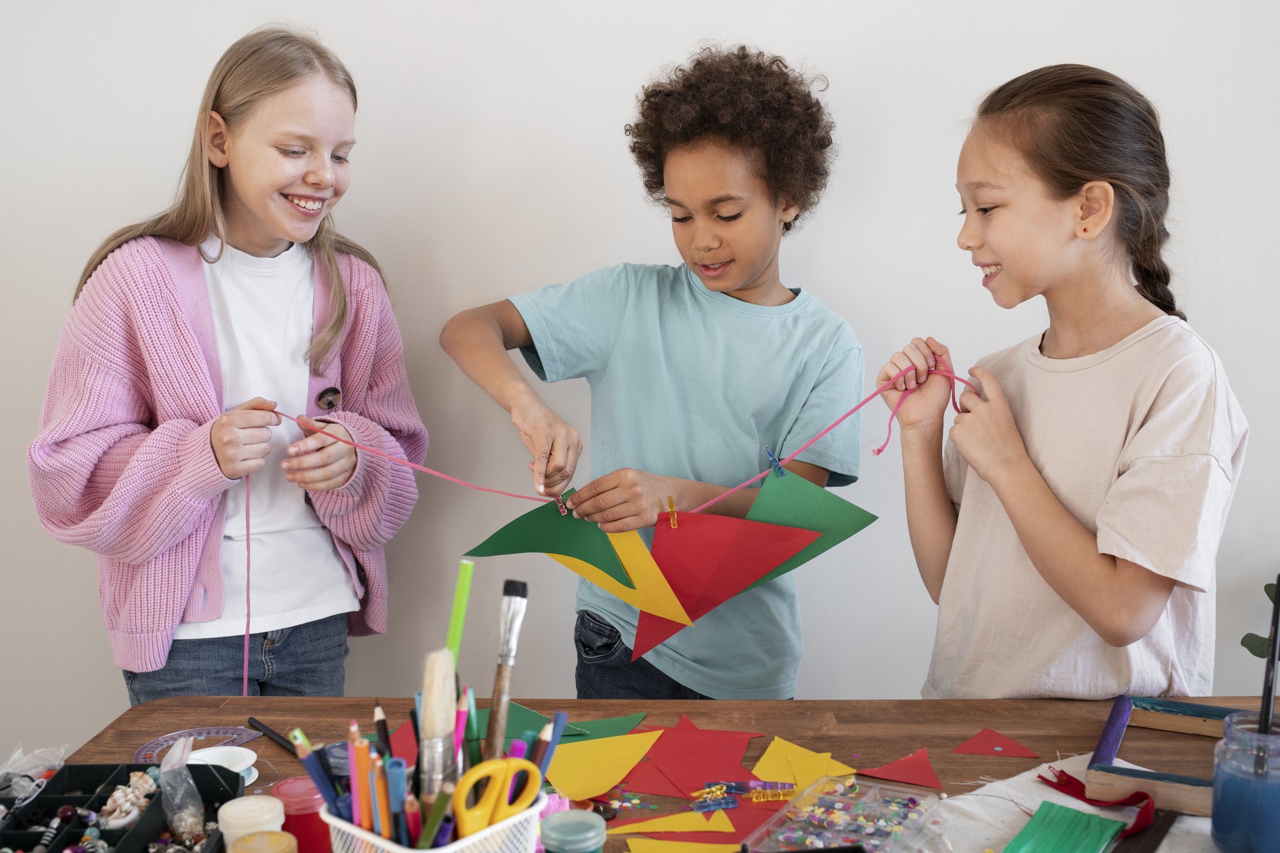 Three children, two girls and a boy, happily connect and hold a colorful paper kite in a craft room with arts and craft supplies on a table.
