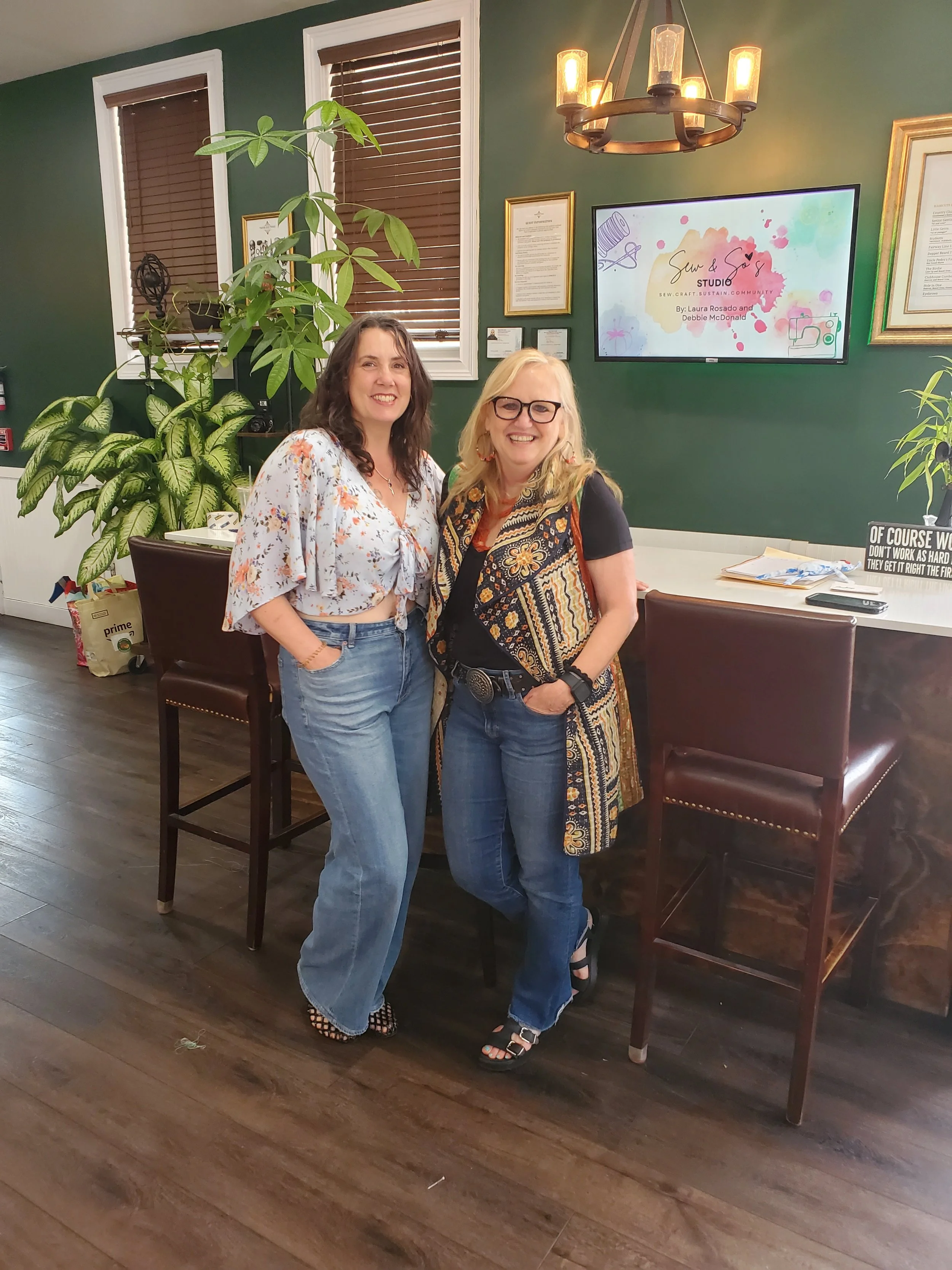 Two women smiling and posing in a room with green walls, wooden floor, and plants. One woman is wearing a floral top and jeans, while the other is wearing a black shirt, jeans, and a patterned vest. There is a television screen in the background displaying the text 'Sun & Sew Studio'.