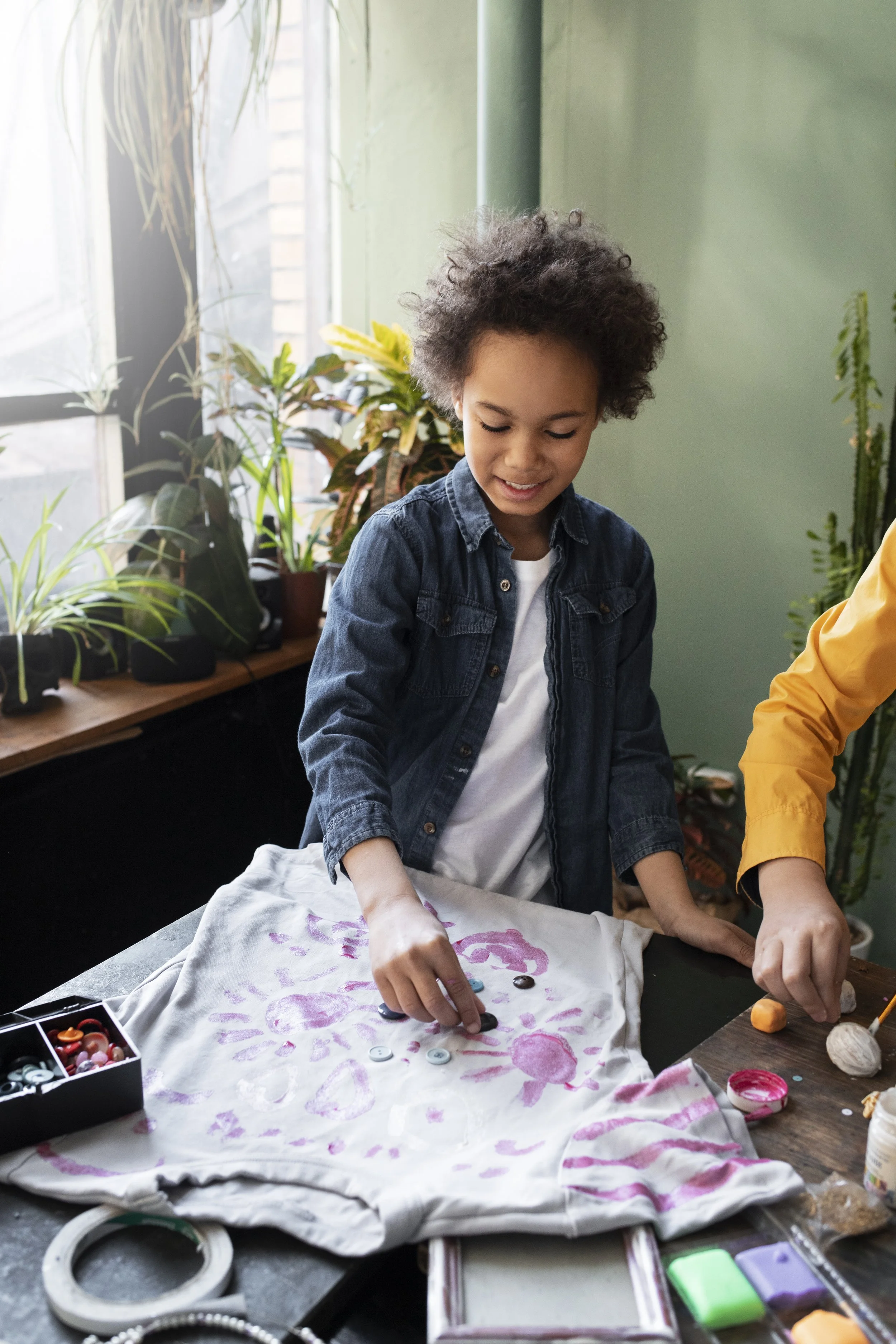 A young boy with curly hair wearing a denim jacket and white shirt decorating a white t-shirt with buttons using fabric paint and stamps, surrounded by arts and crafts supplies on a table.