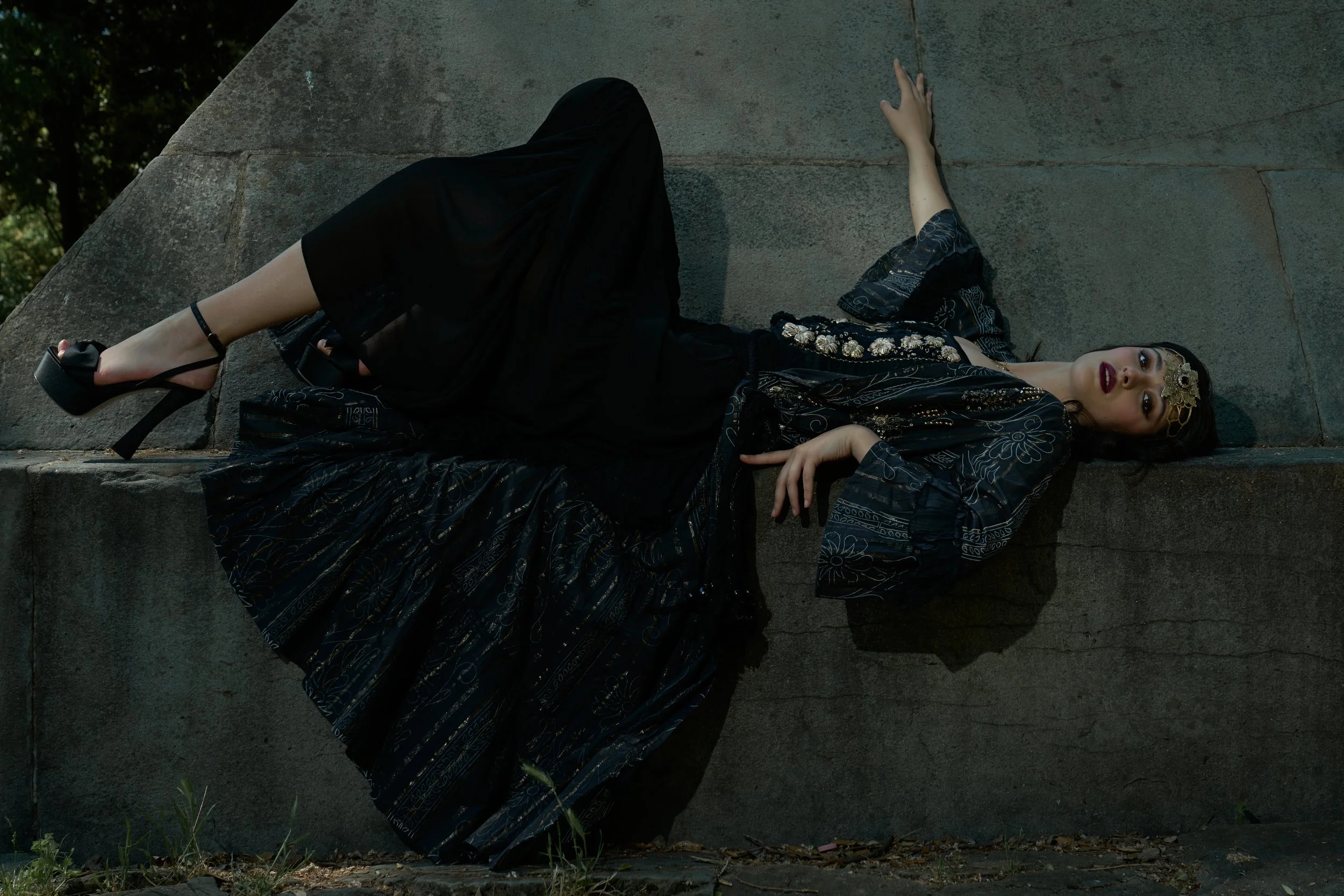 A woman with dark hair and makeup lying on a large concrete structure outdoors, wearing a black embroidered dress, high heels, and a decorative headpiece.