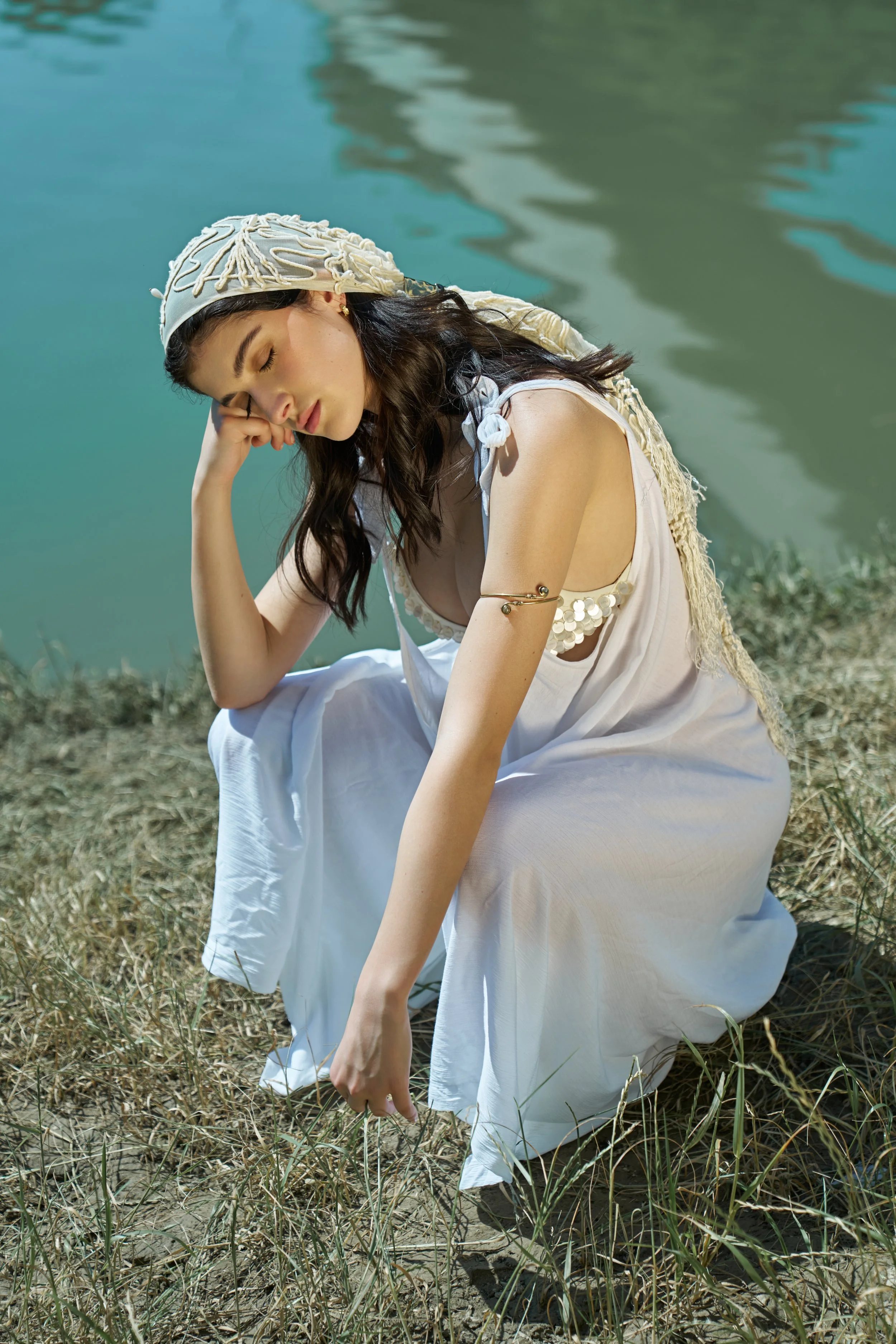 A young woman in a white dress and a beaded headscarf sitting on grass by a body of water, with her eyes closed and her head resting on her hand.