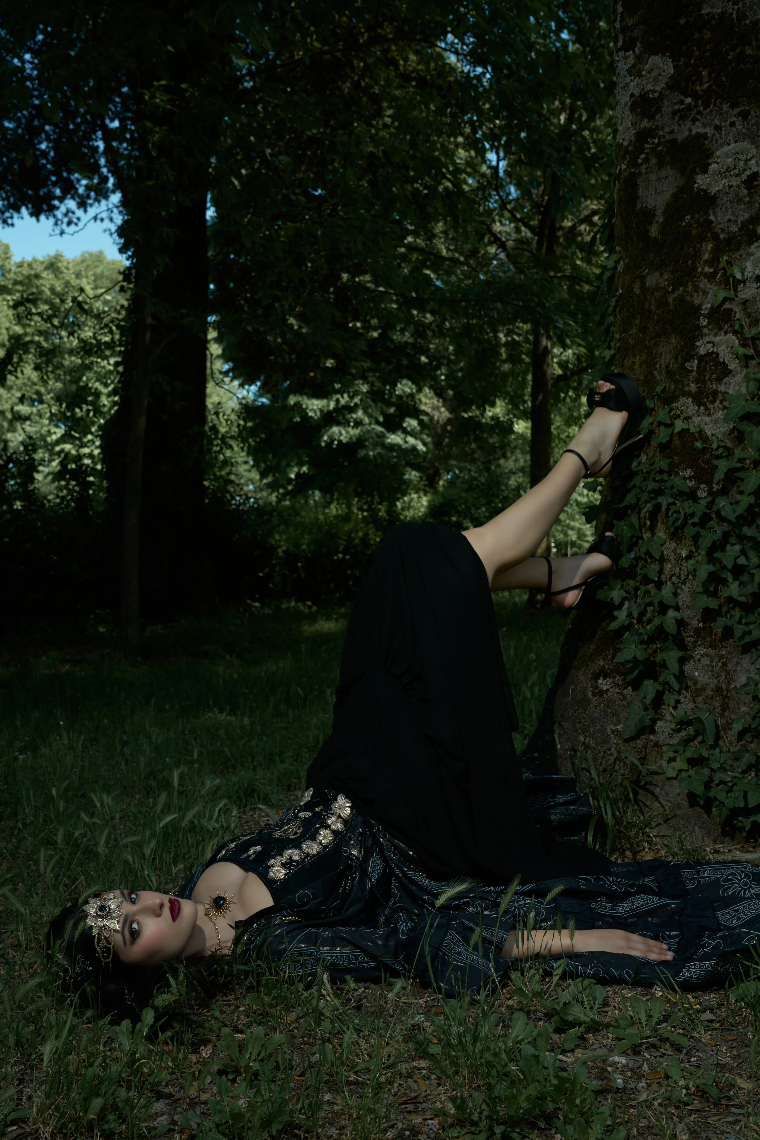 A woman dressed in dark, ornate clothing and jewelry lays on the ground next to a large tree in a forest, with her legs resting against the tree trunk and her upper body and head on the grass.