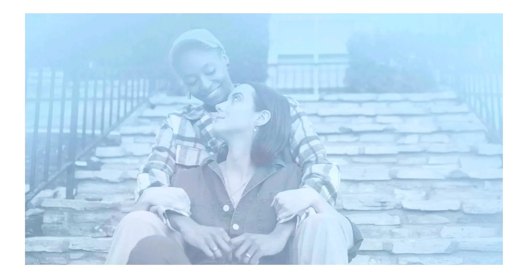 Two women sitting on outdoor stone stairs, one embracing the other, smiling and looking into each other's eyes.