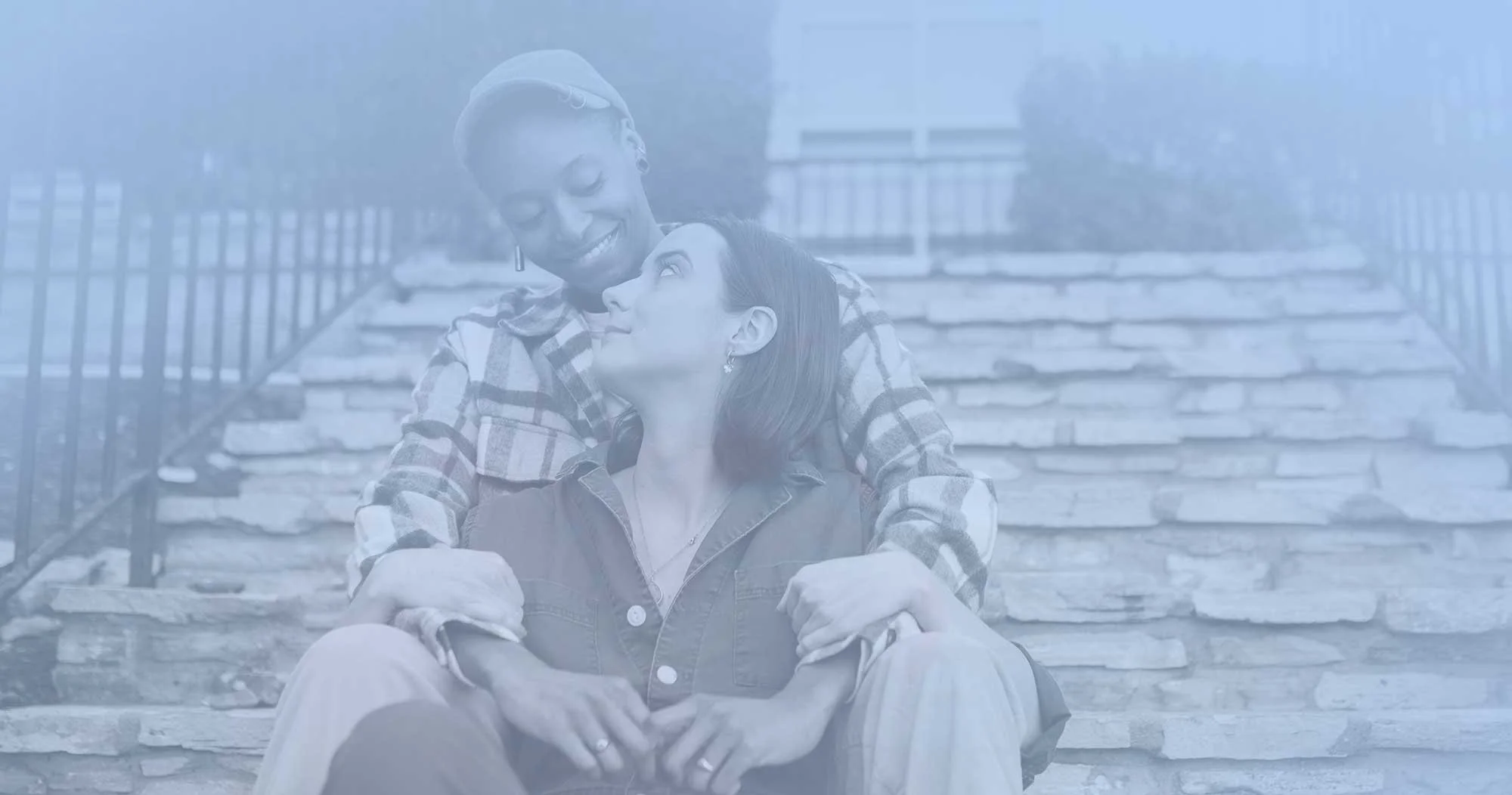 Two women sitting on outdoor stone stairs, one embracing the other, smiling and looking into each other's eyes.