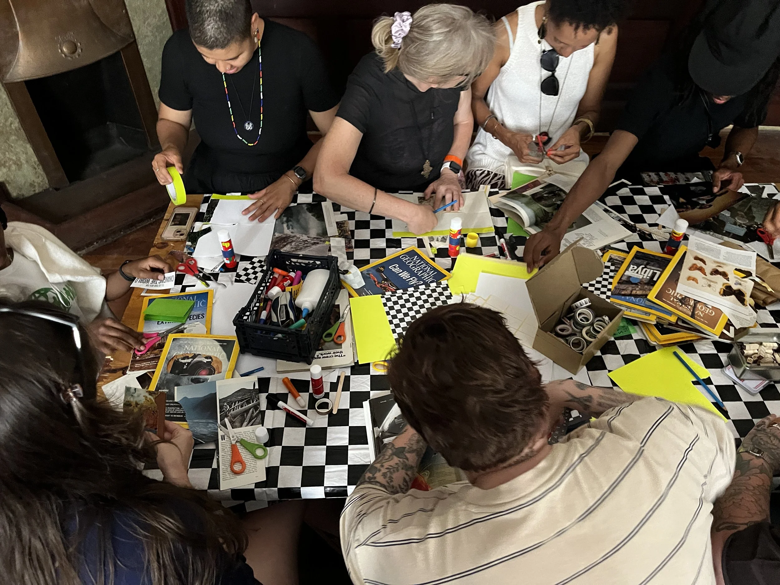 Group of diverse women and men gathered around a table working on craft activities, with magazines, scissors, glue, and markers, in a casual indoor setting.