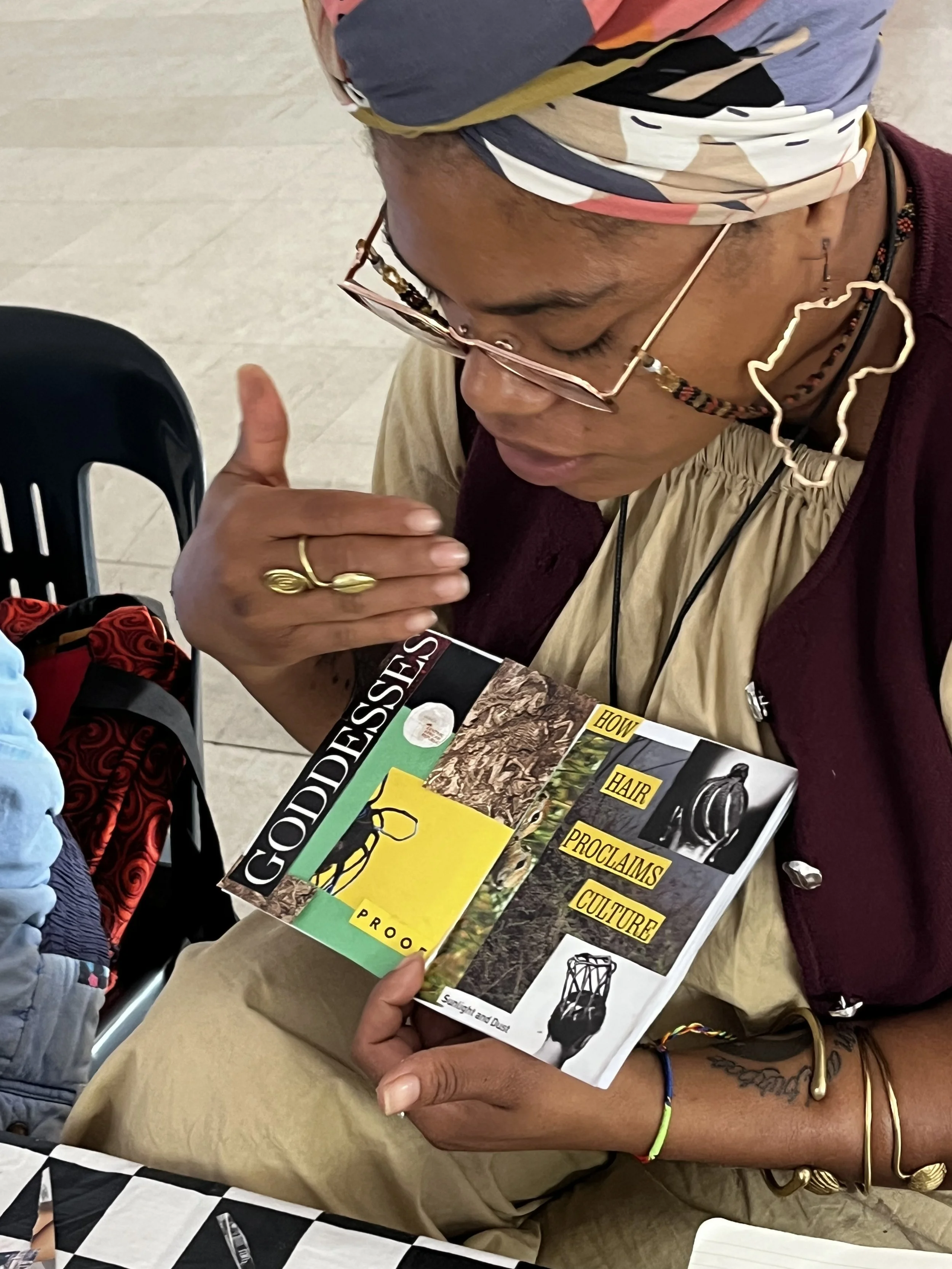 Person holding a brochure about the culture and history of Goddesses, proclaims, and proclamations, with a person sitting nearby, wearing jewelry and a colorful headscarf, at a table with a black-and-white checkered tablecloth.