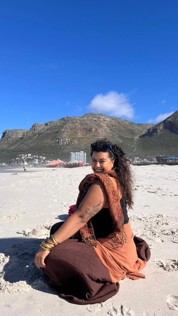 A woman sitting on a sandy beach, smiling at the camera, with mountains and clear blue sky in the background.