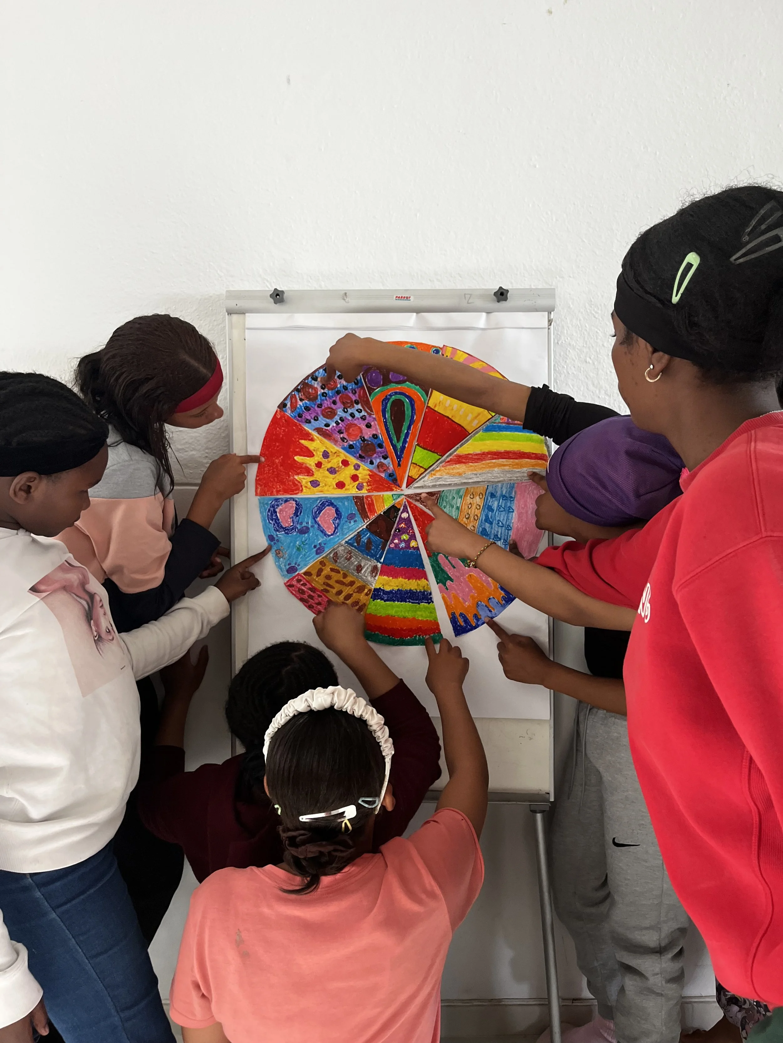 Children and an adult are gathered around a colorful, circular art piece on a whiteboard, pointing and observing it.