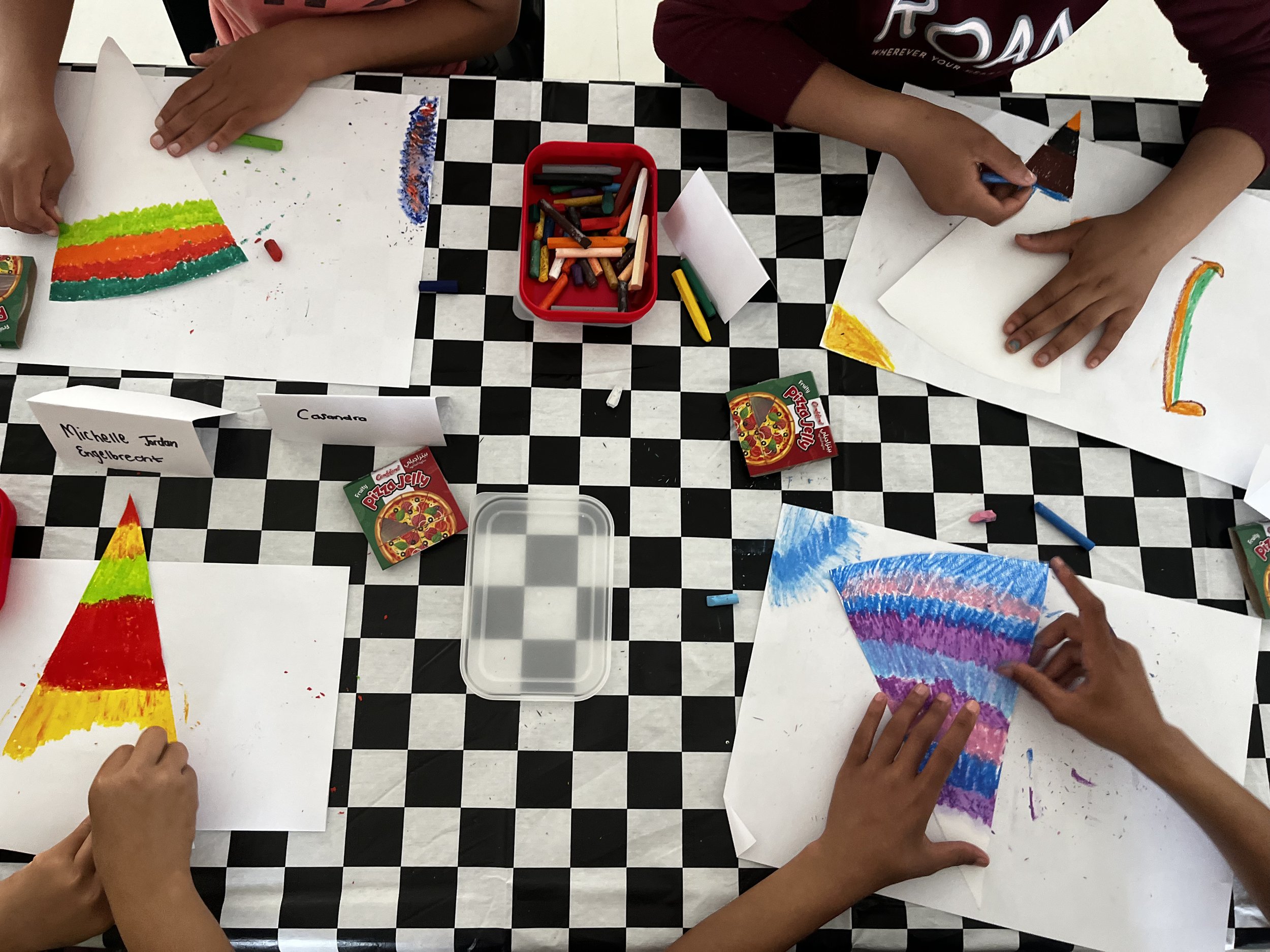 Children drawing colorful rainbow-themed artwork with crayons and pastels on white paper at a checkered table.