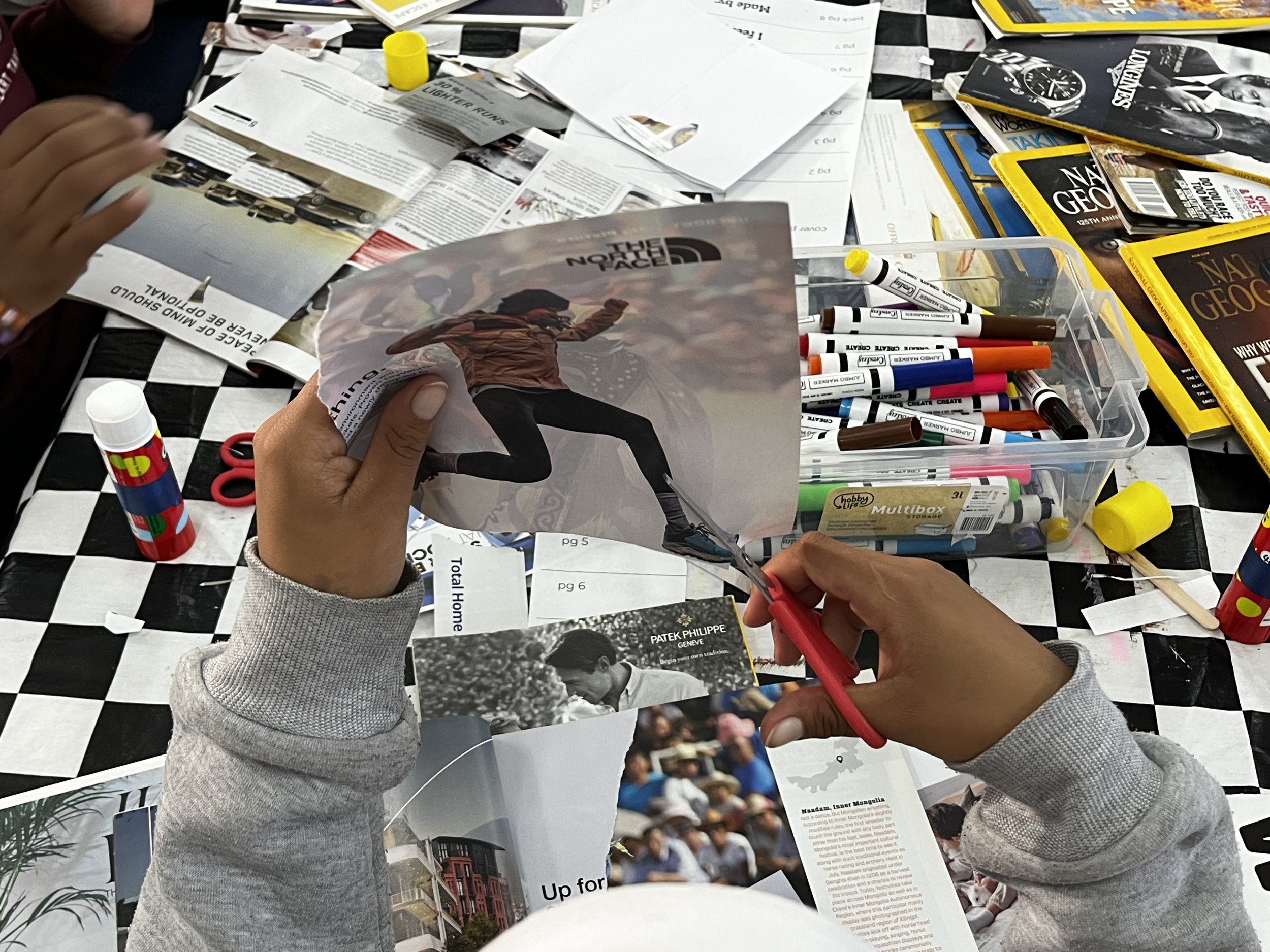 People sitting at a table covered with newspapers, brochures, markers, glue, and scissors, engaging in a craft activity involving cutting and gluing magazine images and articles.