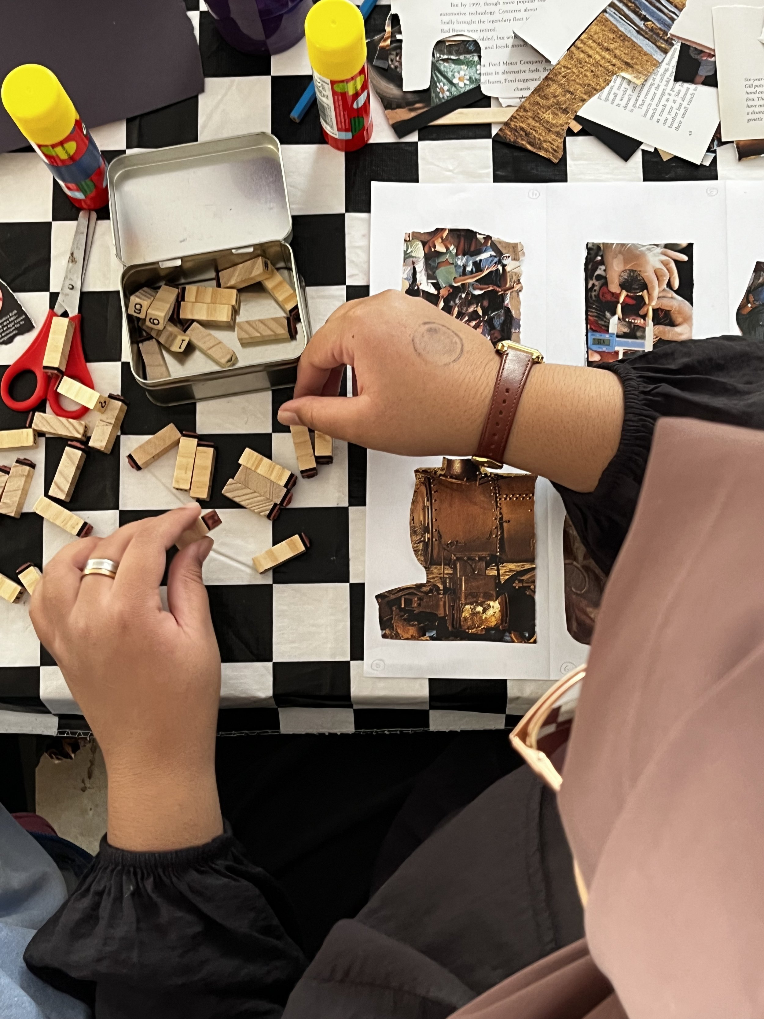 Top-down view of a table with black and white checkered pattern, scattered papers, a pair of red scissors, a metal container with wooden blocks, and bottles of glue. A person with a light skin tone, wearing a leather strap watch, is reaching for the 