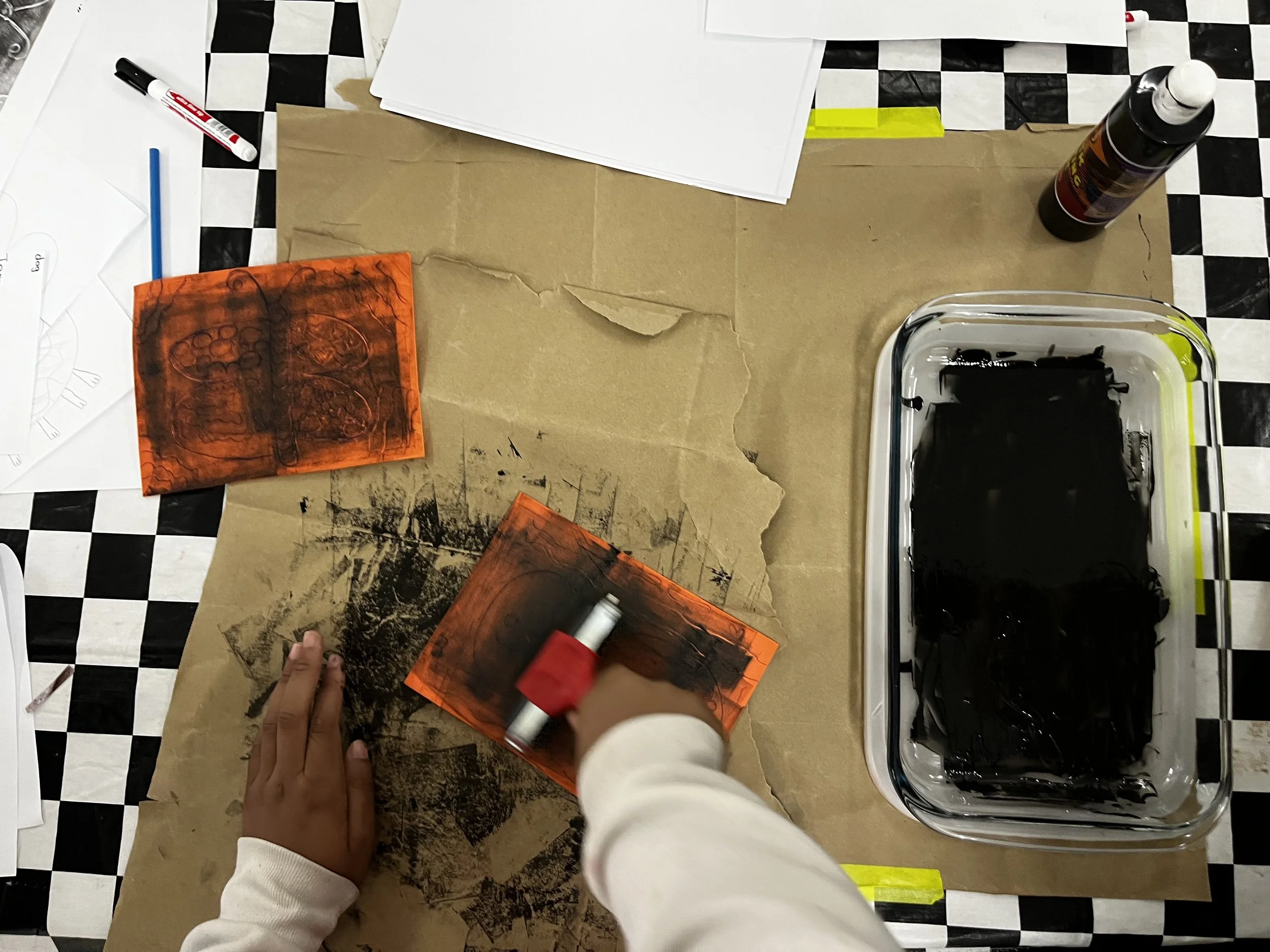 A person spray painting orange on a block of wood at a cluttered workspace with papers, markers, and a black stove.