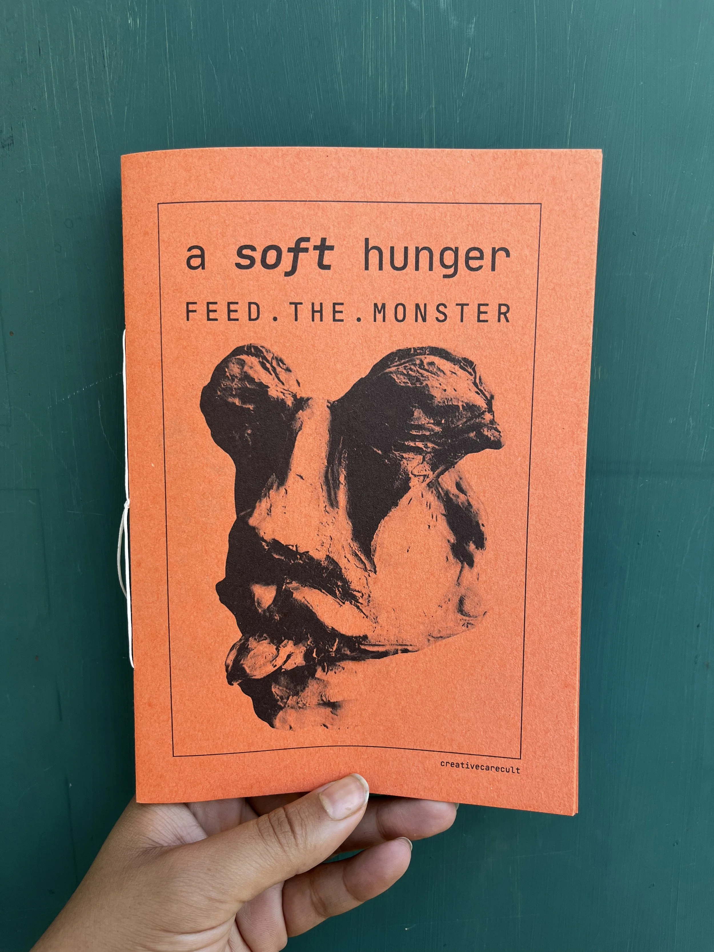 A person holding an orange card featuring black text that says 'a soft hunger FEED THE MONSTER' and an abstract black and brown illustration of a creature's head.