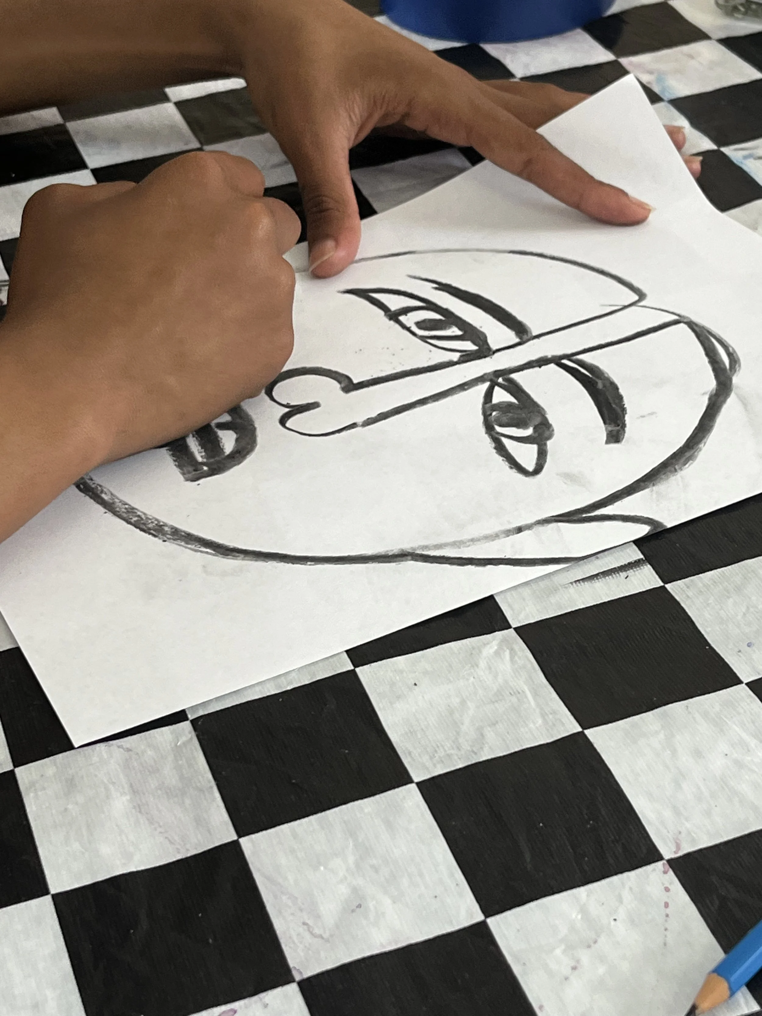 Hands drawing a face on a piece of paper with a black marker, placed on a black-and-white checkered tablecloth.