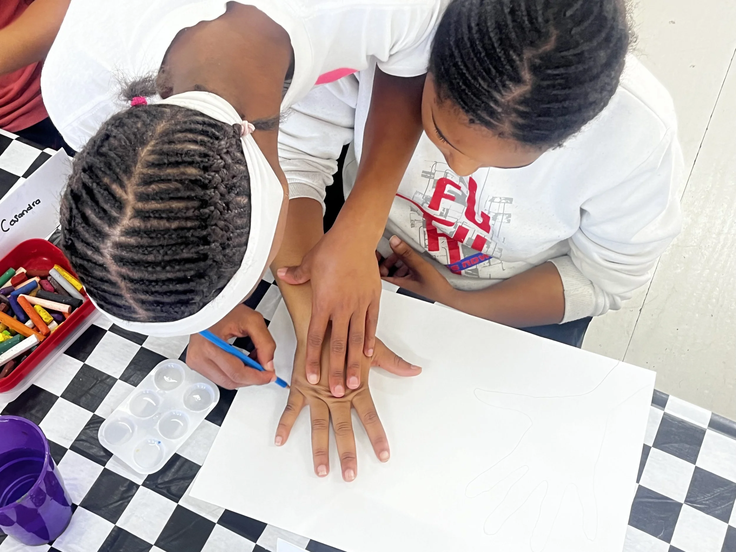 Two children with braided hair engaging in a hands-on activity at a table, with one child's hand on top of the other's, surrounded by colorful markers or crayons, on a black-and-white checkered floor.