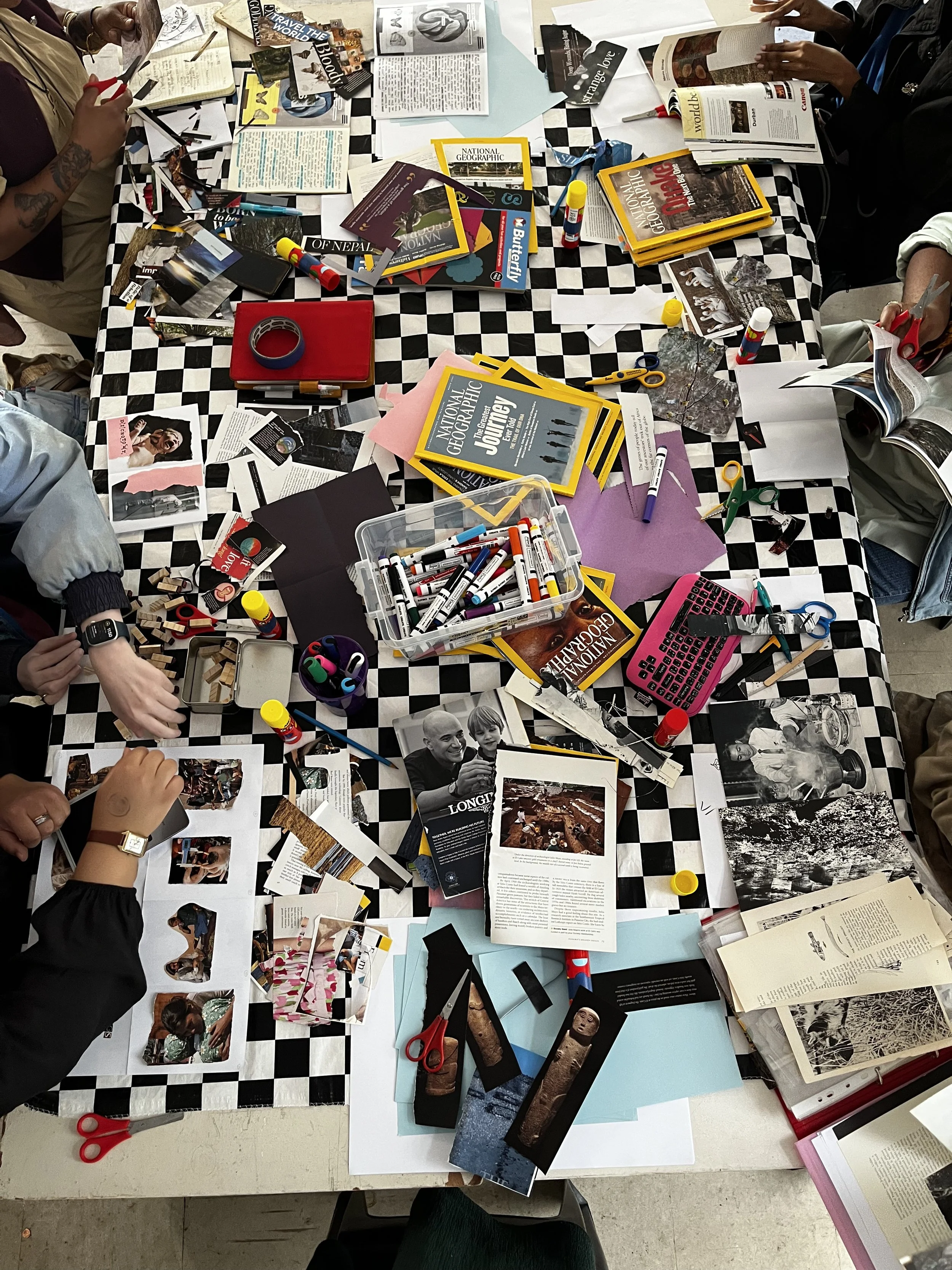 A group of people working on a project at a table covered with photos, magazines, markers, scissors, glue sticks, and papers. The table has a black and white checkered tablecloth.