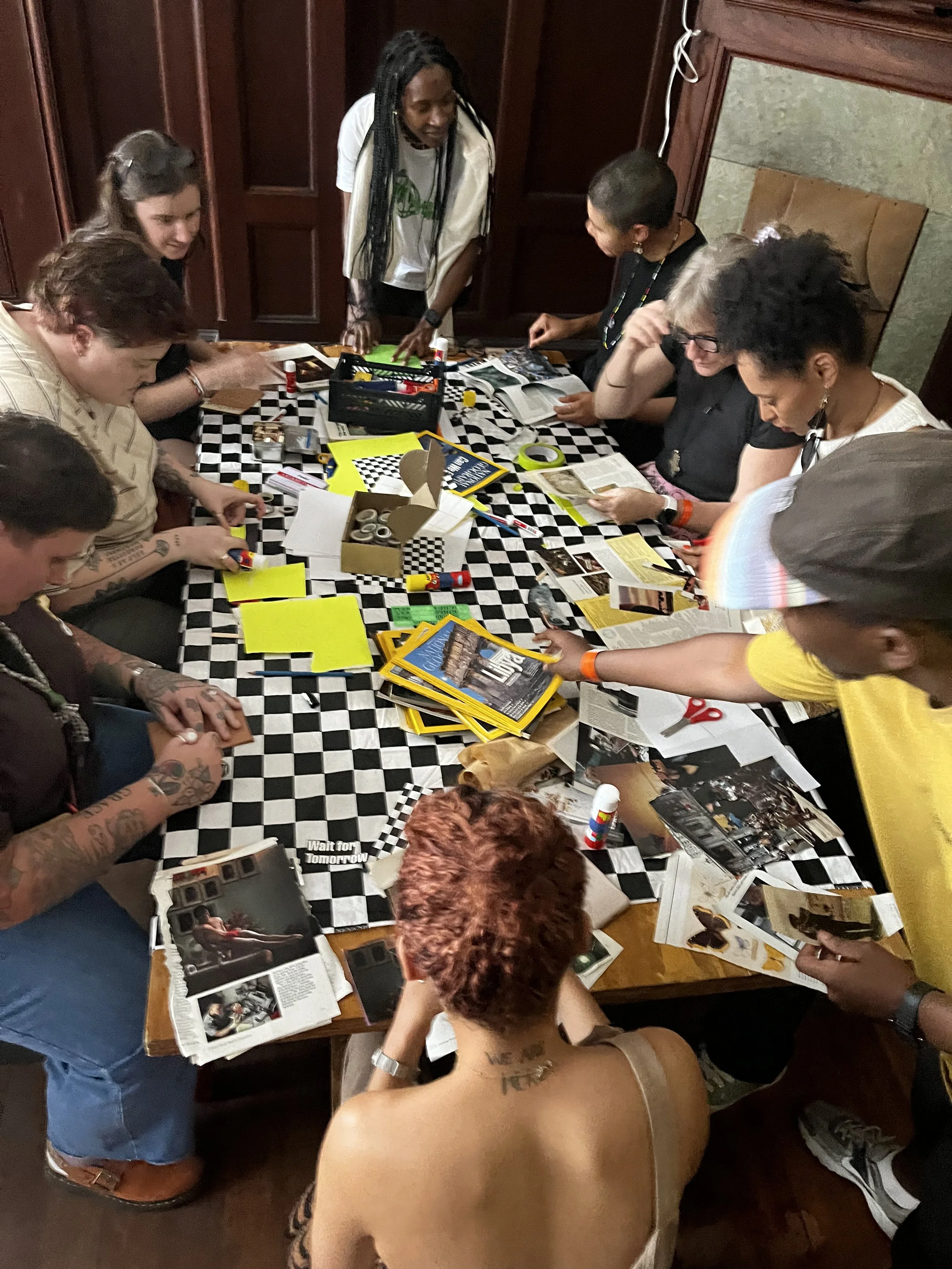 Group of people gathered around a table filled with magazines, photos, scissors, glue sticks, and other craft supplies, apparently working on a collage project.